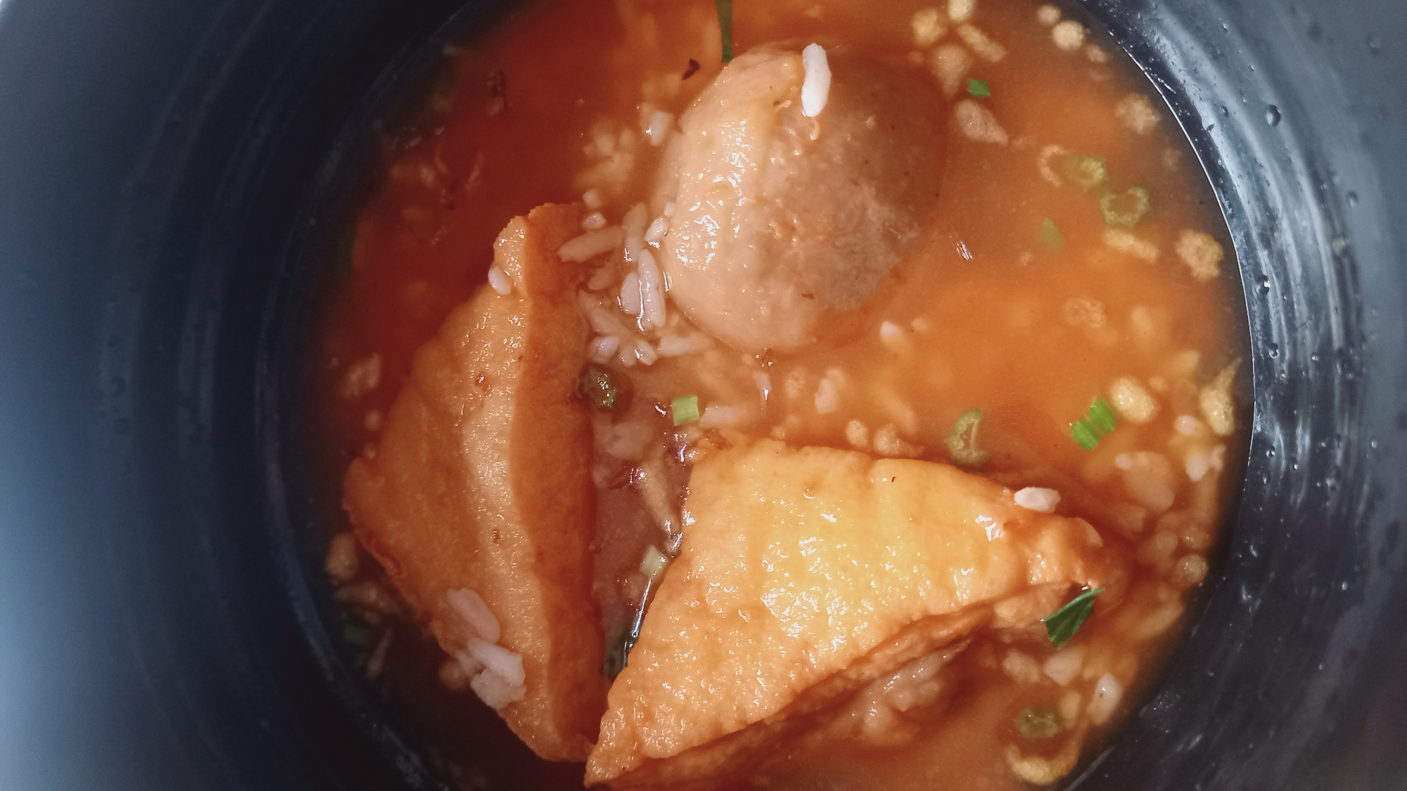 A close-up, overhead shot shows a dark-colored bowl filled with a savory broth. Inside the bowl, there are several pieces of fried tofu, roughly triangular in shape and golden brown. A single, large meatball, light brown and spherical, is also visible. White grains of rice are scattered throughout the broth, along with finely chopped green onions. The broth itself is a reddish-orange color, suggesting a spicy or tomato-based flavor. The texture of the food appears soft and saucy. The bowl's dark, matte finish contrasts with the vibrant colors of the food. The scene is likely indoors, possibly in a home or restaurant setting in Leuwisadeng, Indonesia, where this type of dish might be commonly served.