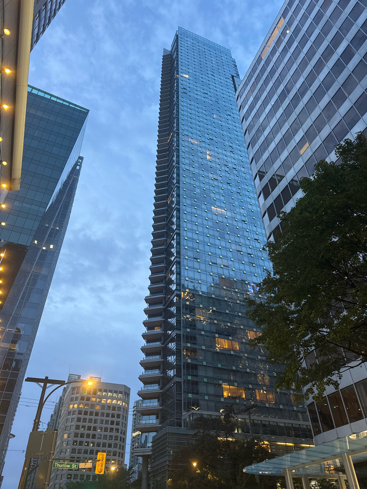 This is a low-angle, wide shot of a city street scene with several modern high-rise buildings in Vancouver, Canada, under a cloudy twilight sky. The prominent structure in the center is a tall, slender skyscraper with a glass facade featuring balconies that ascend its height. To its left is another glass-fronted building with a distinctly angular design, reflecting the sky. To the right of the central skyscraper is a broader, more traditional office building with a facade of numerous square windows, some illuminated with warm light, indicating occupancy. In the foreground, a street sign reads "Thurlow St," and a traffic light is visible. Streetlights and string lights on an overhang add to the urban ambiance. Mature trees with dark green foliage are present, partially obscuring some of the buildings. The overall impression is one of a bustling urban environment at dusk.