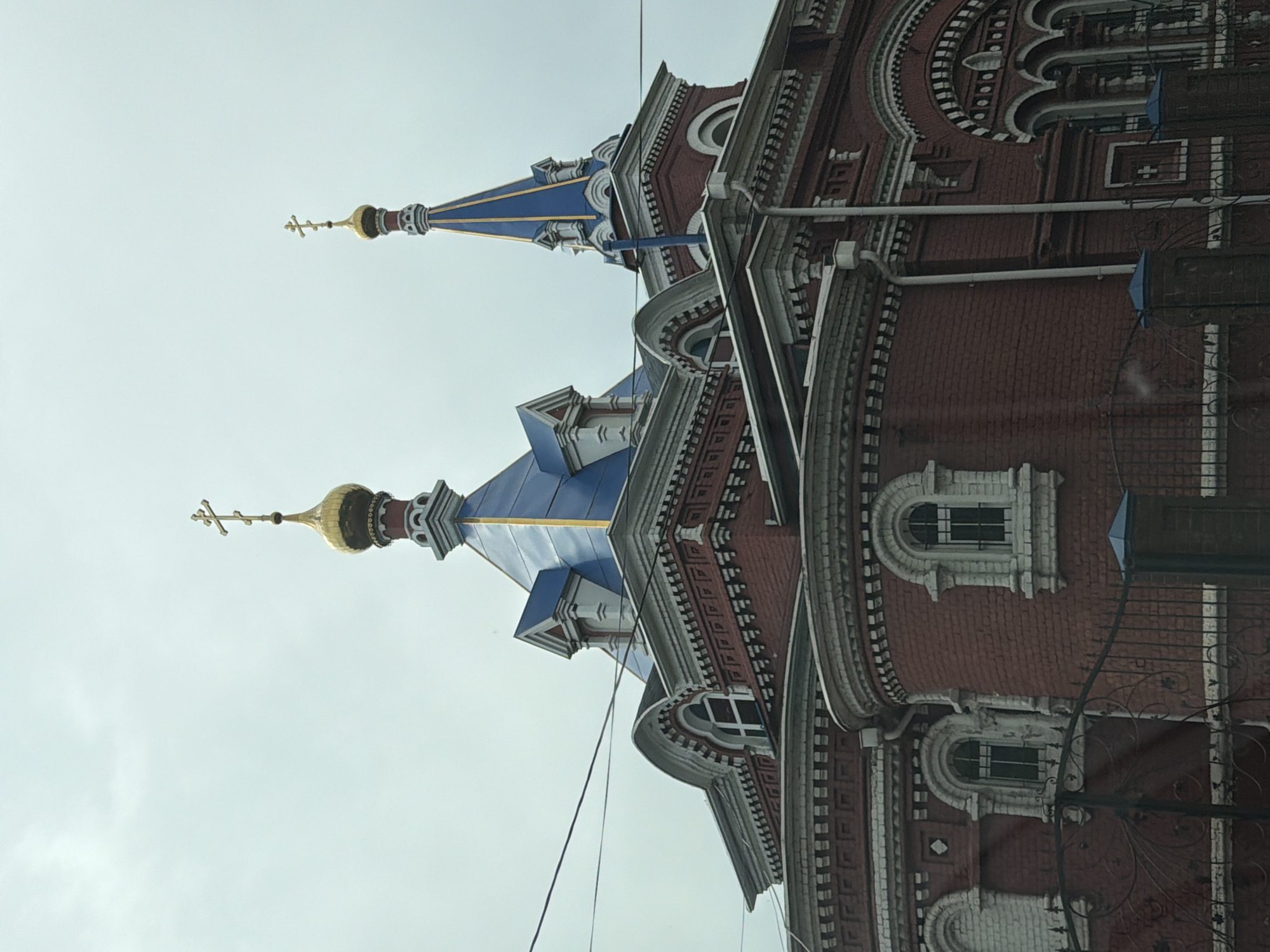 A low-angle view of a brick and stone building with two domes located in Осеченки, Russia. The building has a reddish-brown brick facade with gray stone detailing and ornate architectural elements. There are several arched windows with white frames. Two of the building's domes are visible. Both are blue with gold crosses at their peaks. A thin wire runs across the frame in front of the building. The sky is gray and overcast. A section of a black metal fence is on the right side of the frame.
