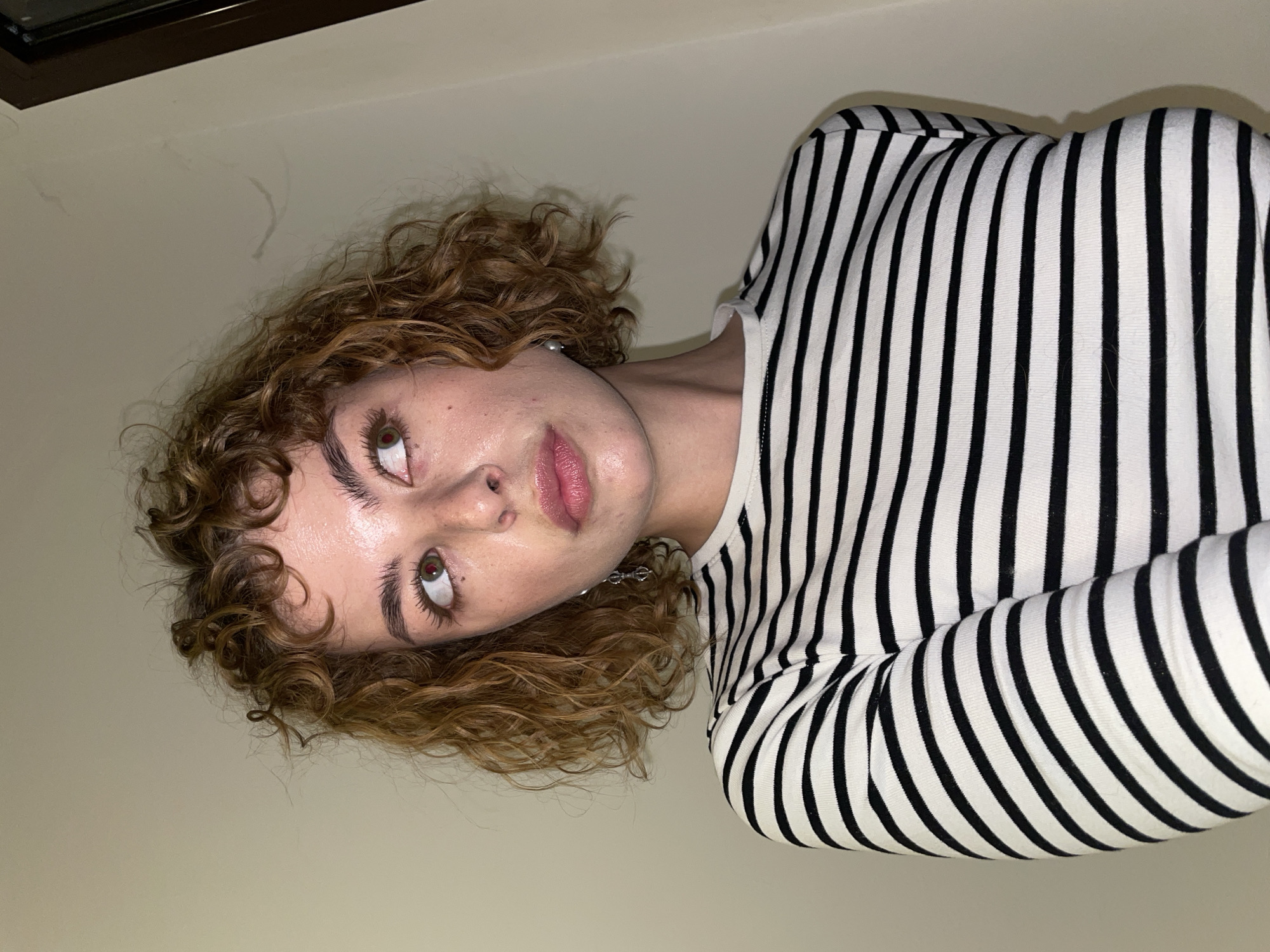 A person with curly, light brown hair is wearing a black and white striped shirt. Their face is tilted upward, and they are looking up and to the left. The background is a light beige wall. The location is not discernible from the image provided.