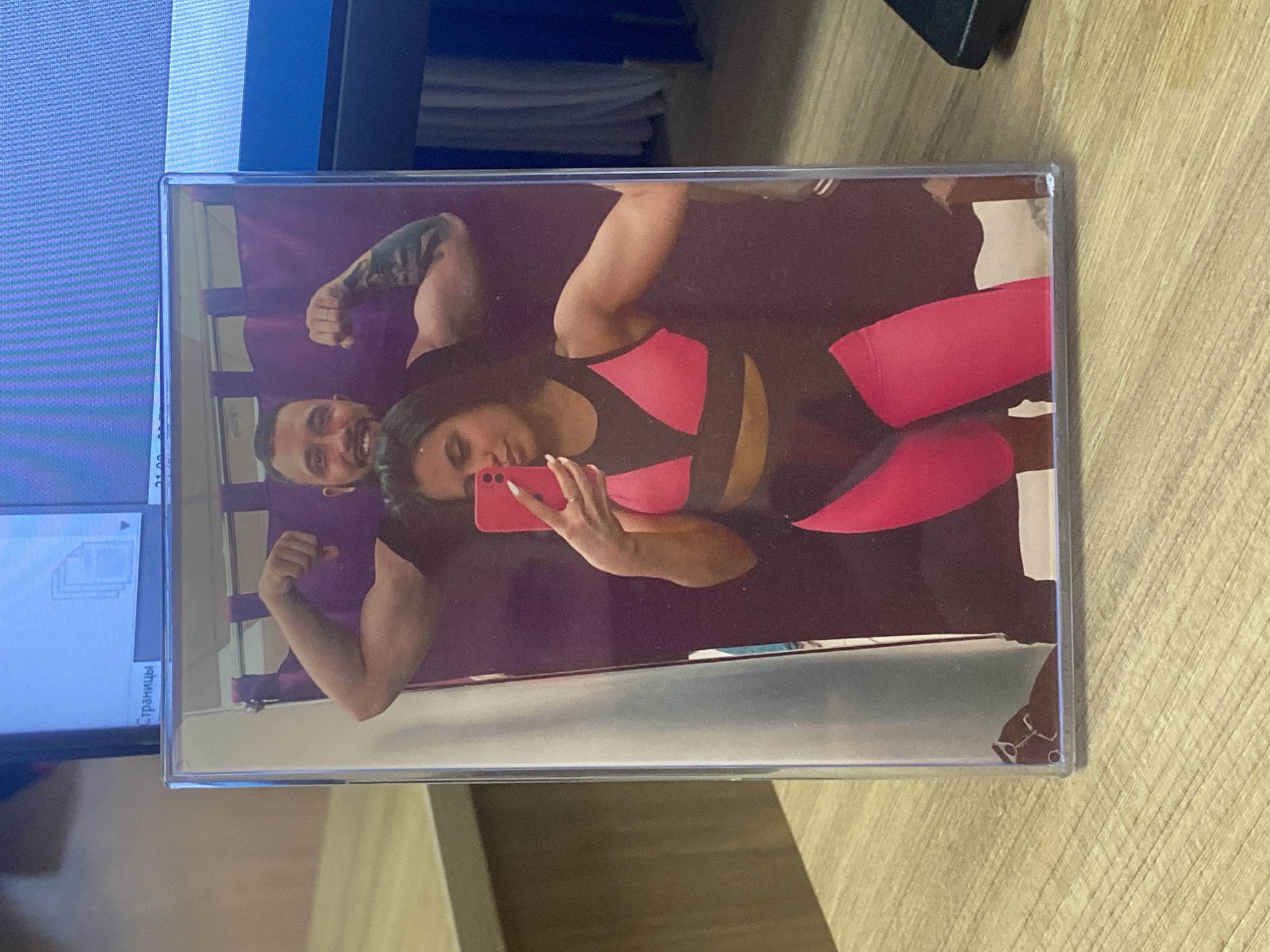 A photo encased in a plastic frame sits on a wooden surface. The photo depicts a couple, a man and a woman, posing in what appears to be a gym or fitness studio. The woman is wearing a pink and black workout outfit and holding a pink smartphone, presumably taking a selfie. The man is behind her, flexing his bicep. The photo is partially reflected in the plastic frame, showing elements of the surrounding environment, including a computer monitor and papers. There is no information to indicate the photo was taken in Luzhitsy, Russia.