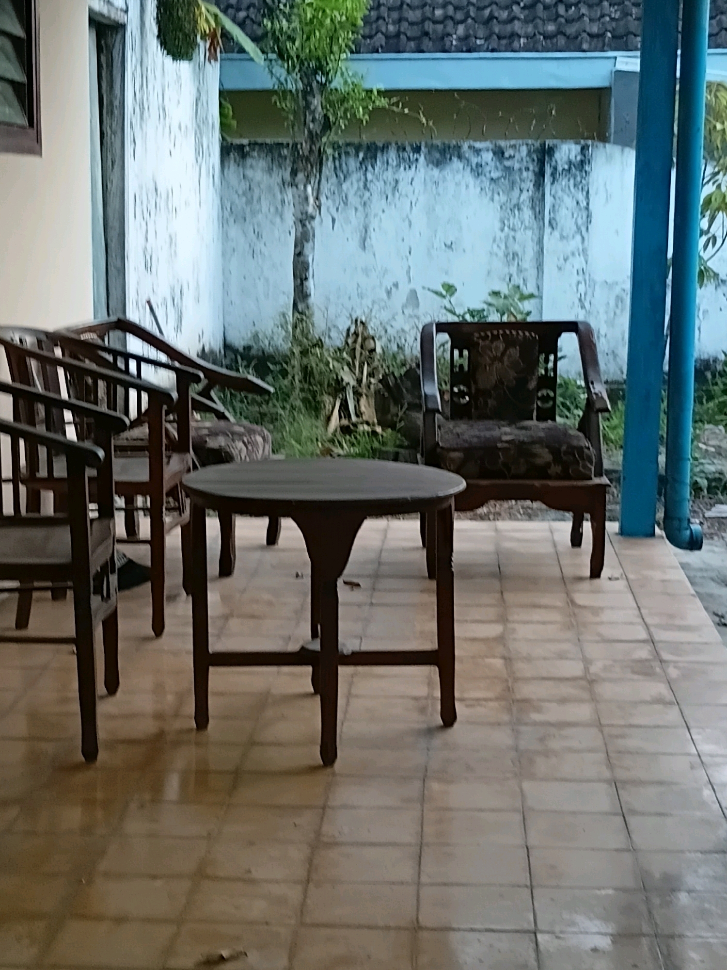 The image depicts a porch area with tiled flooring in Jember, Indonesia. Three wooden chairs and a wooden table are present. One chair is positioned to the left, facing slightly away from the viewer, with another chair partially visible behind it, also angled away. A third, more ornate wooden chair with patterned upholstery is situated to the right, facing the viewer. A dark-colored, round wooden table stands in the center foreground. The background shows a white, textured wall, a tiled roof, and some greenery. A blue downspout is visible on the far right. The scene appears to be outdoors and appears to be uninhabited at the time of capture.