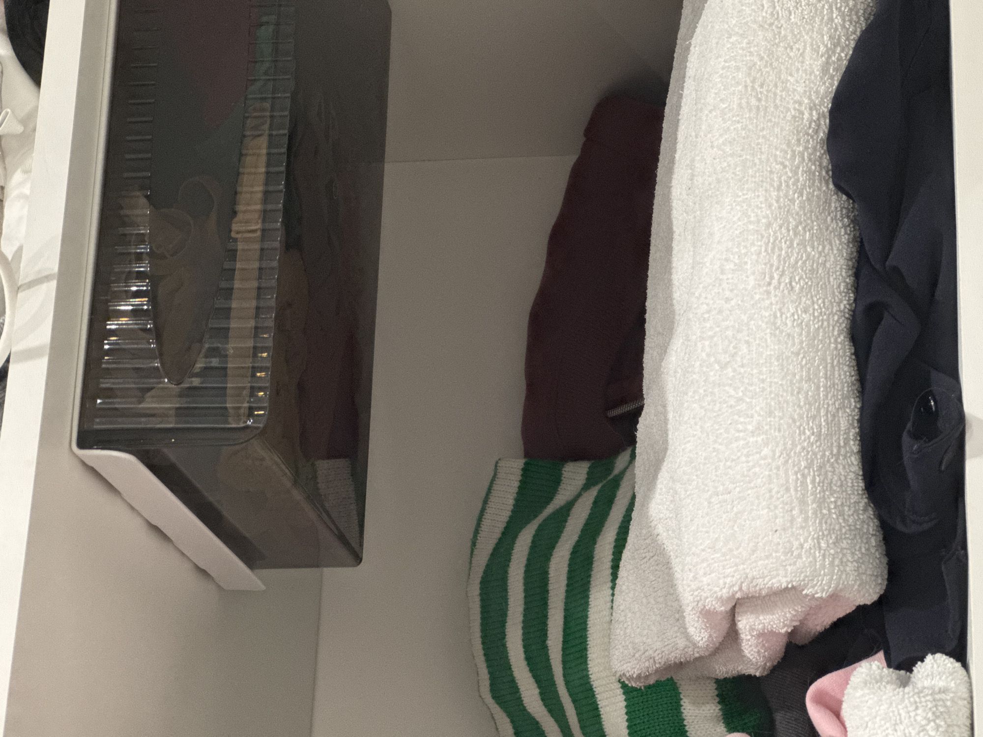 In an indoor environment, a white shelf is visible. On the shelf, a clear plastic drawer with a white frame is placed, partially obscuring folded clothing items beneath it. To the left, a green and white striped sweater is folded, and a white towel is stacked on top of dark blue fabric. A dark red garment is visible behind the sweater. The scene is lit from above, casting shadows.