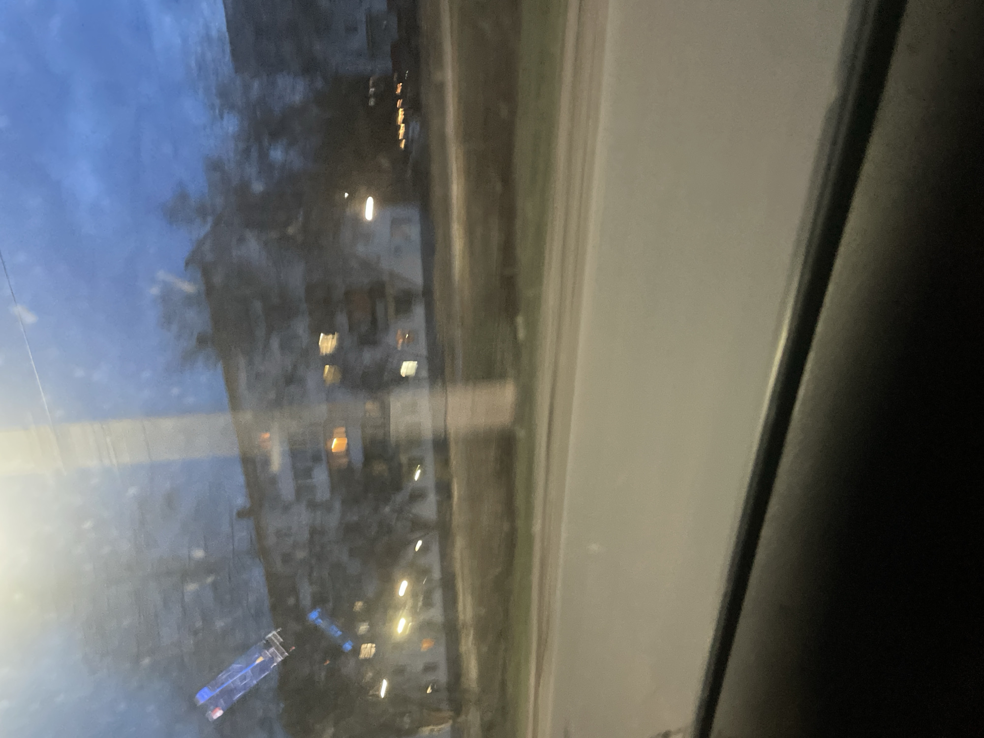 View from a window in Chaikovsky, Russia, at dusk or night. The scene outside is blurred and slightly distorted by the glass. Streetlights and the outlines of buildings are visible in the background. A blue and white object, possibly a vehicle, sits at the bottom center of the view. The window frame is visible on the right side, with a grey wall and a darker element to the right of the window frame.