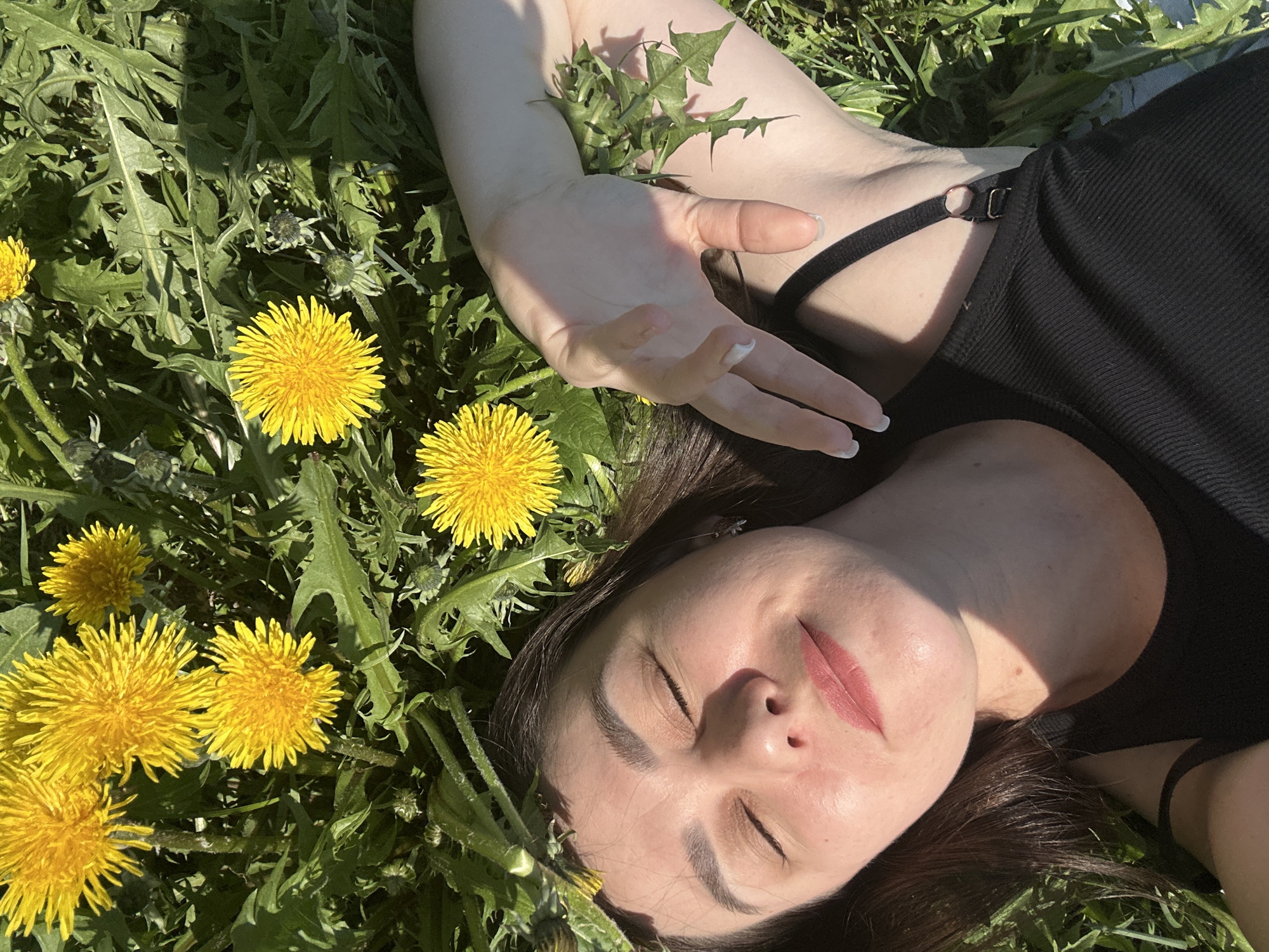 A young woman is lying on her back in a field of dandelions, eyes closed, as if relaxing or sleeping. She has dark hair, fair skin, and is wearing a black top. Her left hand is extended, resting amongst the dandelions. The location is a field in the city of Chaikovsky, Russia. The scene appears to be outdoors on a sunny day.