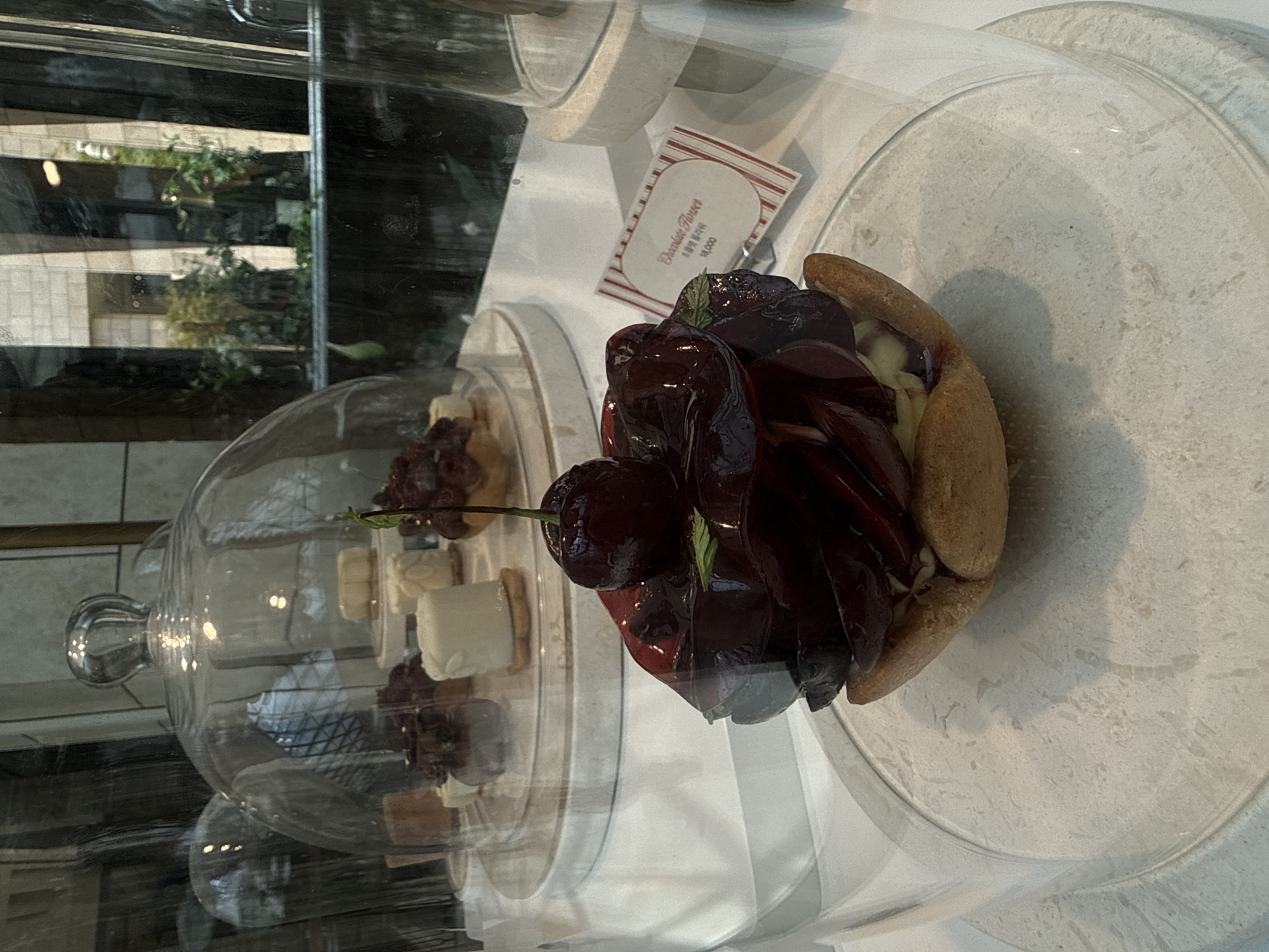 A close-up, eye-level shot captures a cherry tart presented under a glass dome in a bakery in Incheon, South Korea. The tart features a flaky crust filled with cream and topped with dark, glossy cherries and a sprig of mint. A small, striped card with "Chocolate Flower" and Korean text is placed near the tart. In the background, other pastries are visible under similar glass domes, and through the shop window, a blurred view of greenery and buildings can be seen. The lighting is soft, highlighting the shine on the cherries.