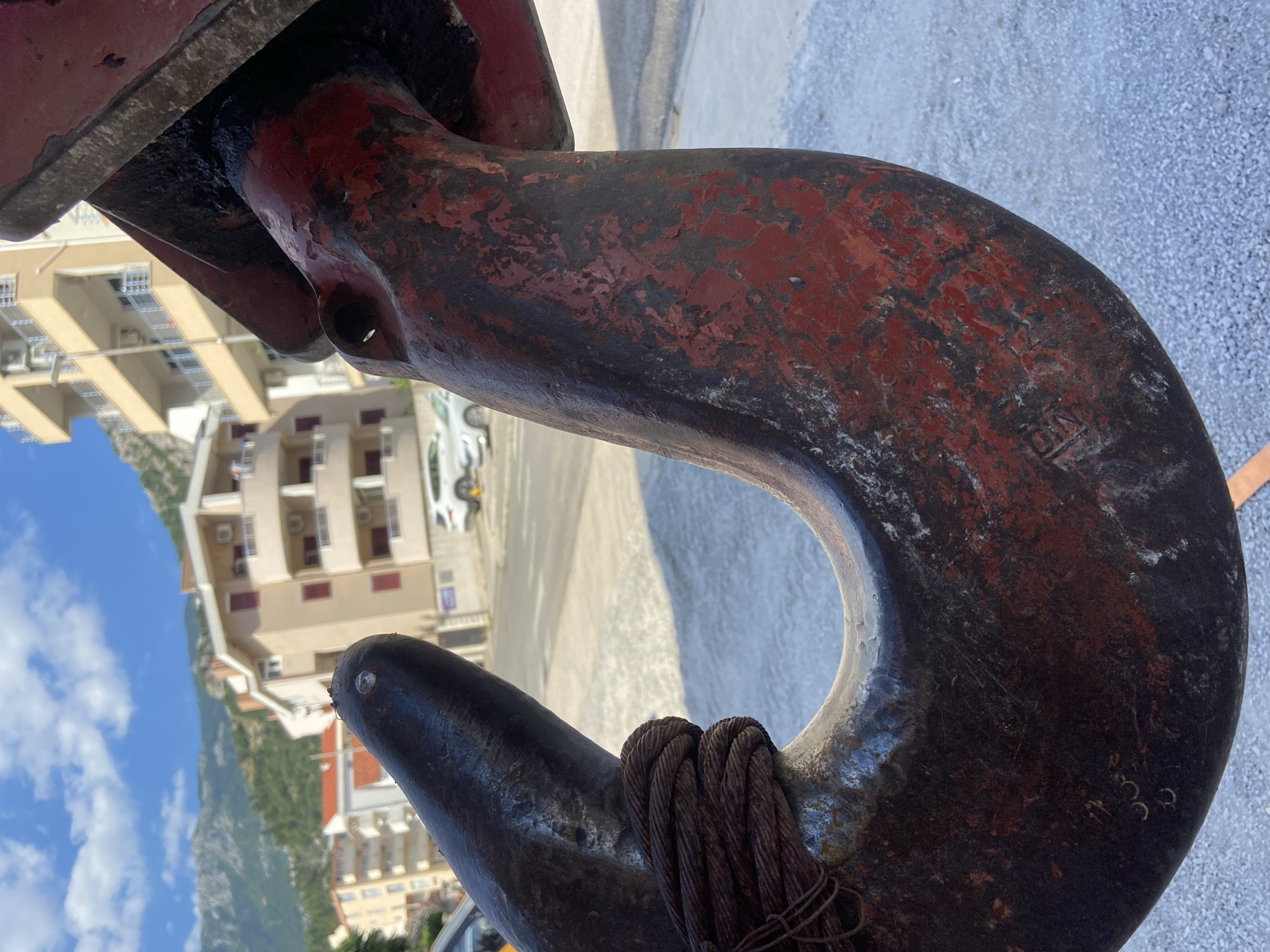 The image is a close-up, low-angle shot of a large, weathered, red and black metal hook, likely part of a crane or heavy lifting equipment. The hook is heavily worn, with significant rust and chipped paint visible across its surface. A thick, multi-strand wire rope is wrapped around the upper part of the hook. The background is out of focus, showing a street scene with buildings and mountains in the distance, characteristic of Budva, Montenegro. The sky is blue with scattered white clouds. The overall impression is one of industrial or maritime equipment against a backdrop of a coastal town.