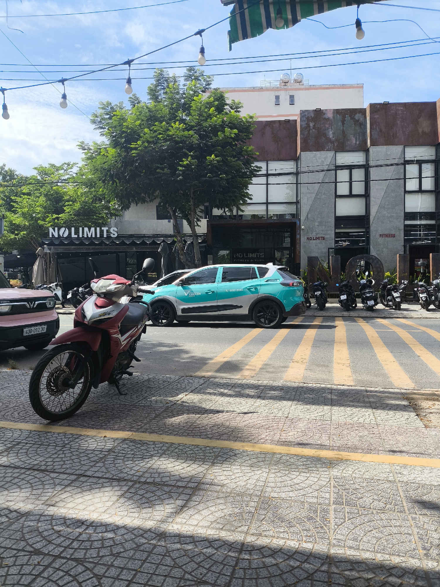 In Đà Nẵng, Vietnam, a street scene is captured on a sunny day. A red and white Honda motorcycle is parked on a pedestrian walkway paved with gray tiles featuring a circular pattern. Adjacent to the walkway is a street with yellow and gray striped crosswalk markings. In the background, a teal and white car, identified by the "XANH SM" livery and "1900 2055" phone number, is parked on the street. Further back, a building facade with large windows is visible, displaying signage that reads "NO LIMITS" and "FITNESS." Another building, slightly behind and to the left, also features the "NO LIMITS" logo over its entrance. Several motorcycles are parked in front of the building. Overhead, a string of bare light bulbs hangs across the street, and a striped awning is visible at the top of the frame. The sky is blue with scattered clouds, and green trees line the street.