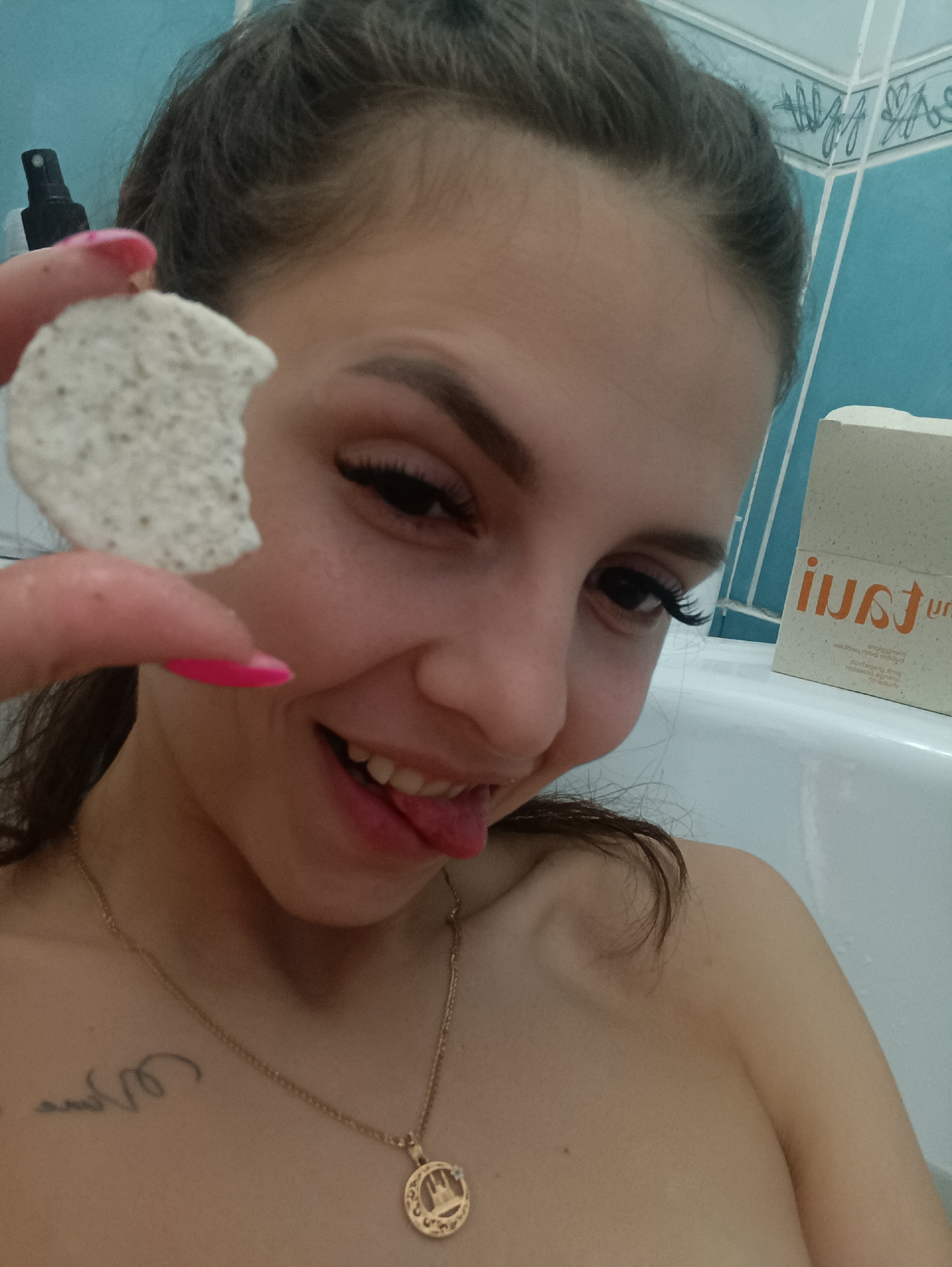 A young woman is taking a selfie in a bathroom. She is fair-skinned with dark hair pulled back. She is sticking her tongue out and holding a round, porous object in front of her face, near her left eye. Her fingernails are painted bright pink. She is wearing a gold necklace with a circular pendant. A tattoo is visible on her chest. The background shows light blue and white tiled walls and a white bathtub. A white and tan box is visible on the edge of the tub, with text written on it. The location is Unknown, Unknown.