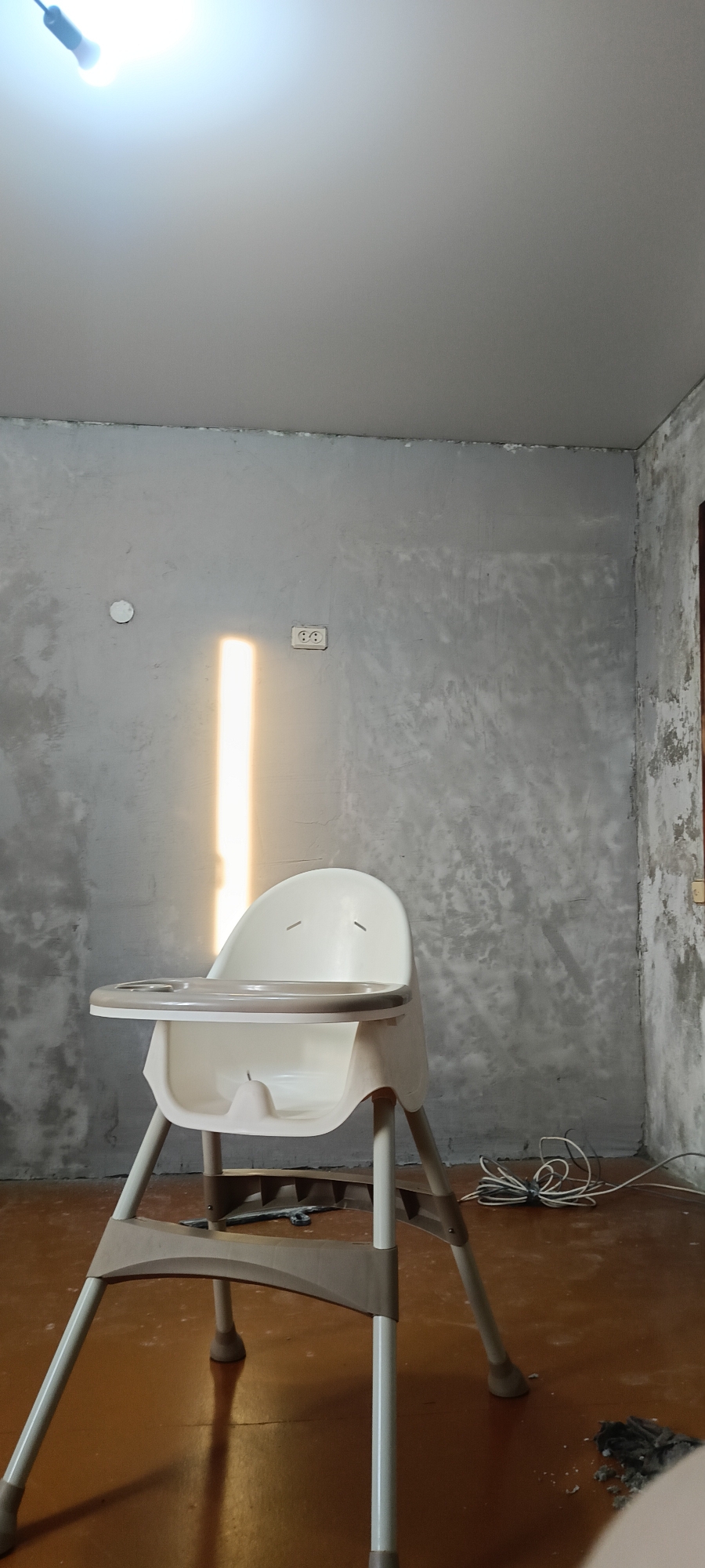 The image depicts an interior scene, possibly a room under renovation or with unfinished walls. The walls are a textured gray, showing variations in color and surface. A bright vertical light source is casting a strong, narrow beam of light across the center of the room. In the foreground, a white and beige high chair is positioned centrally. The floor is a reddish-brown color, with some debris visible, particularly in the lower right corner. Electrical cords are coiled on the floor near the base of the chair. A lightbulb is visible in the upper left corner, illuminating the ceiling. There are no people visible in the image. The setting appears to be a residential interior, possibly in the city of Хохлы, Russia.