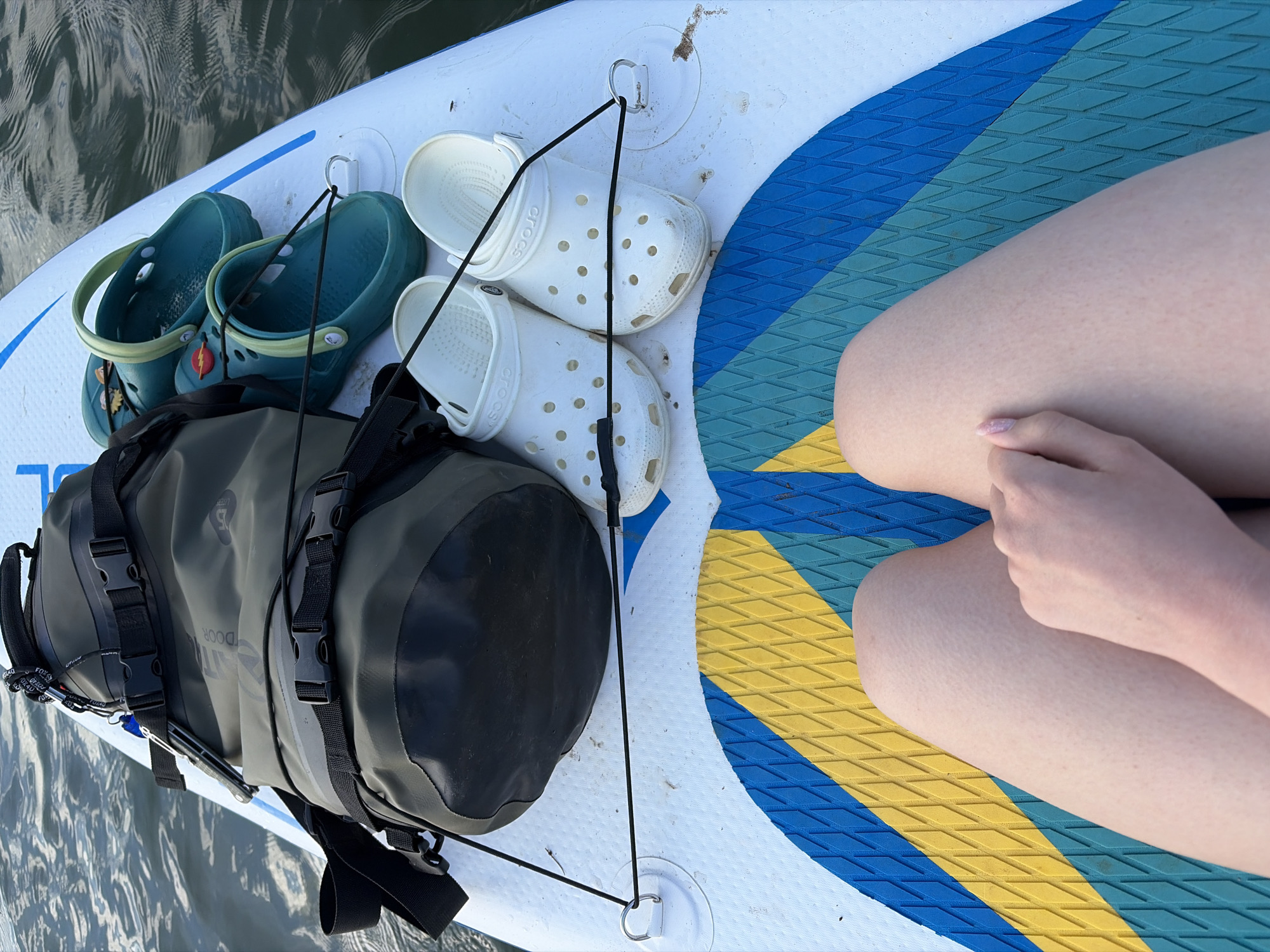The image shows a top-down view of a paddleboard floating on the water, presumably in a body of water near Selychka, Russia. On the paddleboard are several items. There is a black and olive green waterproof bag secured with black bungee cords to attachment points on the board. Next to the bag are two pairs of Crocs: one pair is teal with yellow accents, and the other is white. To the right of the Crocs are a person's thighs and hand resting on the board. The board itself has a blue and yellow patterned non-slip surface. Water surrounds the paddleboard.