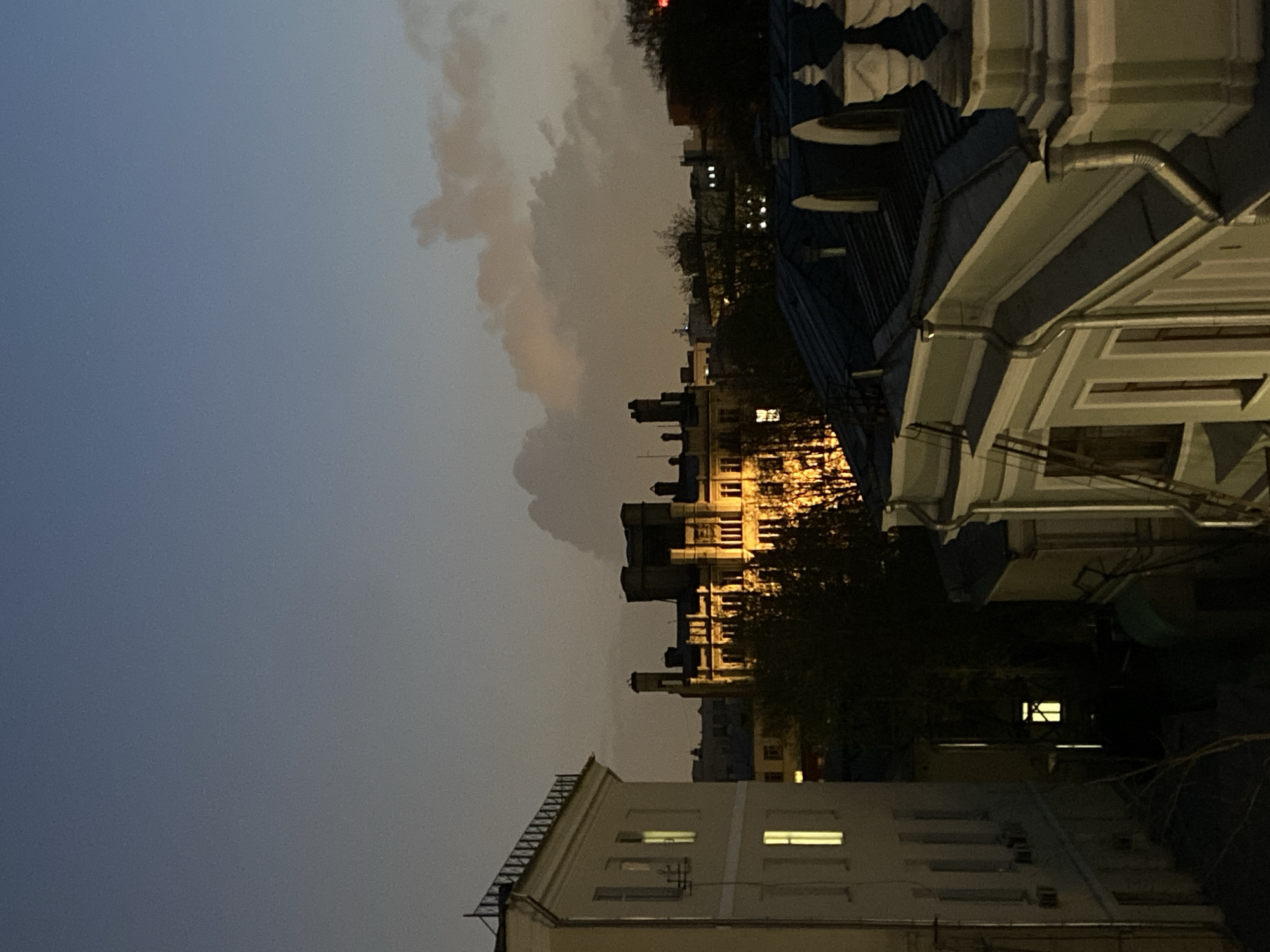 The image captures an urban cityscape at twilight or early night, providing a view across rooftops and buildings under a cloudy sky. The overall mood is quiet and atmospheric, with the warm glow of artificial lights contrasting with the cool, dim natural light.
In the immediate foreground, towards the right side of the frame, is the architectural detail of a building, likely the one from which the photo was taken. It features decorative white cornices and dark rooflines, with a prominent metal downspout running vertically down the structure.
Across the midground, a cluster of buildings with numerous brightly lit windows stands out, casting a warm yellow light. These buildings vary in style and height, creating an uneven skyline. Partially bare trees are visible between the structures, their branches silhouetted against the illuminated facades. Another cream-colored building with some illuminated windows is visible in the lower-left corner of the frame.
The background is dominated by the expansive sky, which shows a gradient from a deep, dusky grey-blue at the top to a lighter, more diffused brownish-orange hue closer to the horizon, suggesting either the last vestiges of sunset or the reflection of city lights on the clouds. Distinct, textured cloud formations are visible against the twilight.
No specific activity or event is captured, but the illuminated windows suggest inhabitants within the buildings. There are no visible people or discernible text. The scene depicts a typical evening in a densely built-up area, consistent with a major city like Moscow, Russia.