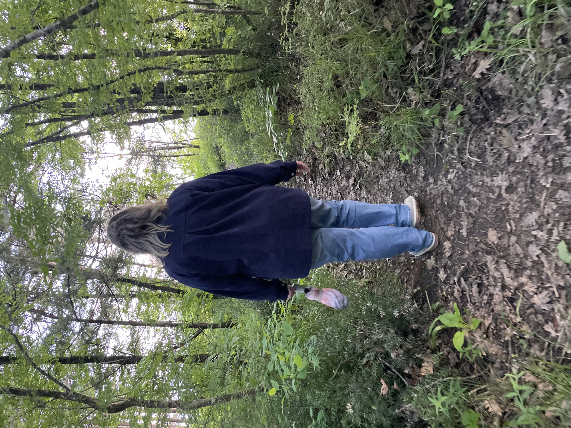 A person wearing a navy blue jacket and blue jeans is walking on a path in a wooded area. The path appears to be covered in dirt and leaves. The trees on either side of the path are green and leafy, creating a canopy overhead. The person is carrying a small, colorful object in one hand. The location is Olginka, Russia.