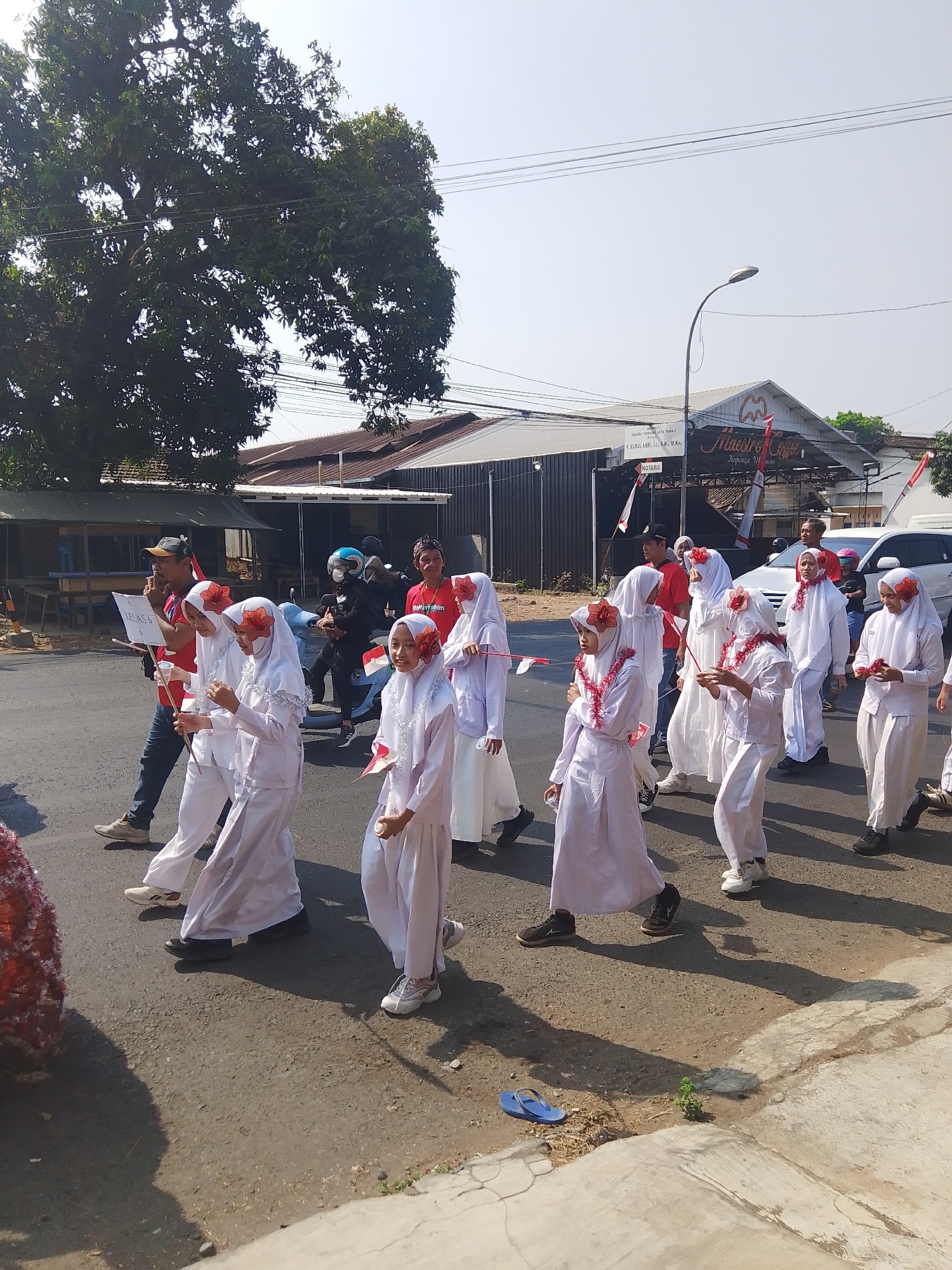 In Kalinyamatan, Indonesia, a group of children dressed in white, long robes and headscarves participate in a parade. Several of the children wear red flowers on their headscarves and around their necks. They carry small Indonesian flags. A man in a red shirt and a baseball cap walks with the children, holding a white sign that says "KELAS 6" (which translates to "CLASS 6"). Other individuals, including adults and some on motorcycles, are also present on the street. The parade takes place on a paved road with buildings and trees visible in the background. The sky is clear and blue, indicating a sunny day.