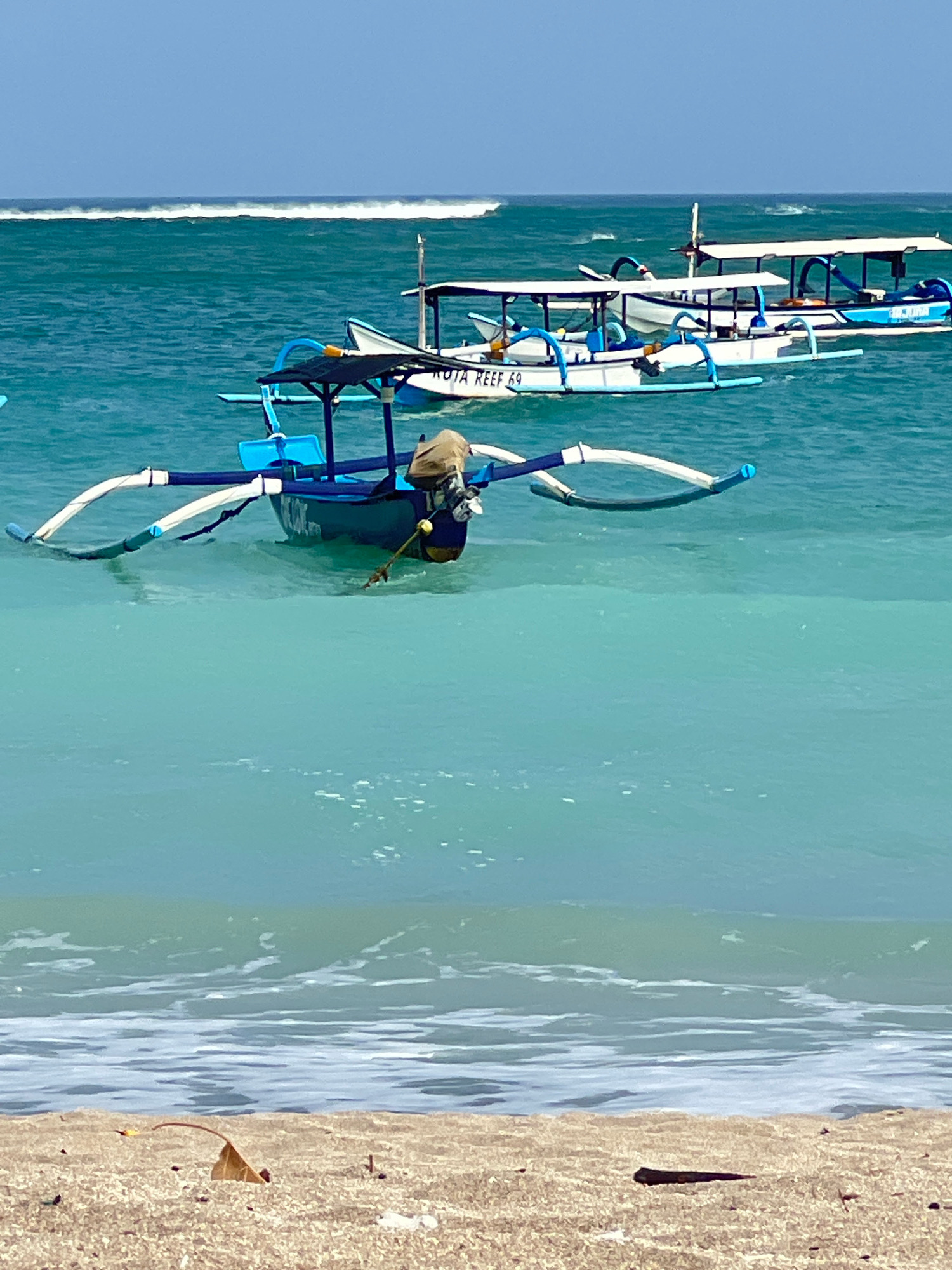 The image captures a serene scene on the coast of Kuta, Indonesia, under a clear blue sky. In the foreground, a sandy beach stretches out, sparsely scattered with debris, including a dried leaf and a dark, elongated object. Beyond the beach, the turquoise waters of the ocean shimmer, with gentle waves lapping the shore. Further out, several traditional Balinese fishing boats, known as jukungs, are anchored. These boats are characterized by their outriggers, which provide stability in the water. The nearest jukung is prominently displayed, its blue hull partially submerged. It features a simple canopy for shade, and a coiled rope extends from its side. In the background, more jukungs are visible, lined up and seemingly waiting for passengers or a fishing trip. The vast expanse of the ocean stretches to the horizon, where a thin line of white surf indicates distant waves breaking. The overall impression is one of a tranquil day in a popular tourist destination, with the iconic fishing boats dotting the beautiful coastal waters of Kuta.