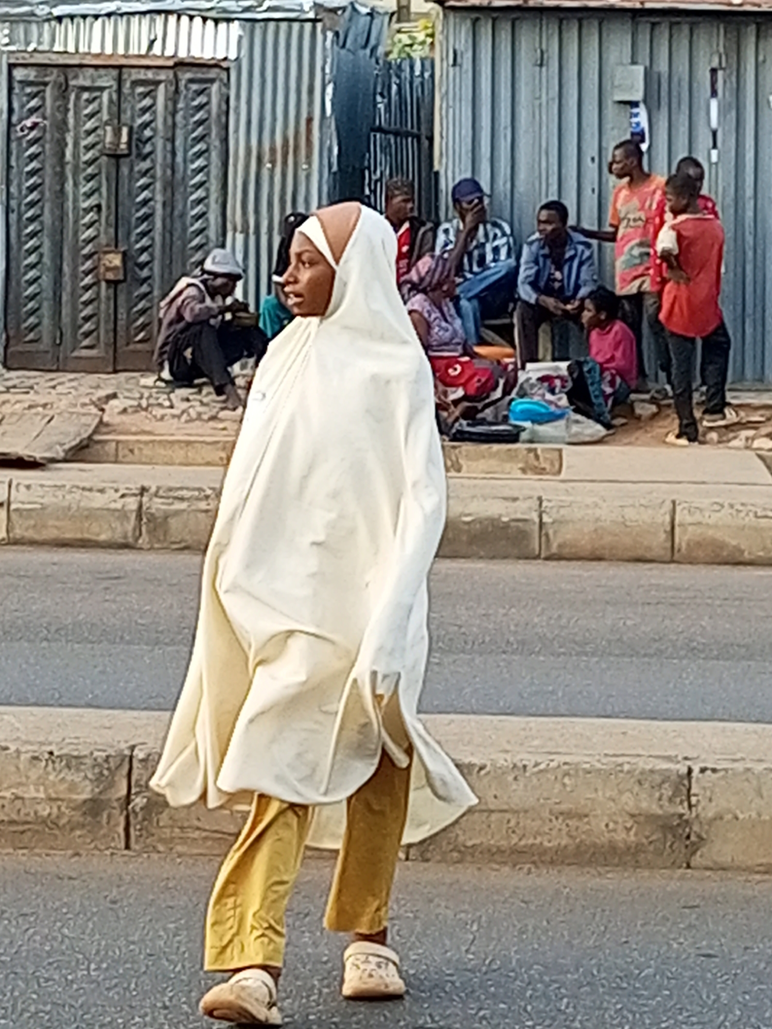 A woman in a cream-colored hijab and gold pants walks across a street in Tempure, Nigeria. She is positioned in the foreground of the image, facing left in profile. In the background, several people are gathered on a sidewalk. To the far left, a man wearing a grey hat sits with his back to the camera. Further to the right, a group of men and women are visible, some seated and others standing. The environment appears to be a street with a corrugated metal building in the background. The ground is paved and a concrete curb separates the sidewalk from the street.