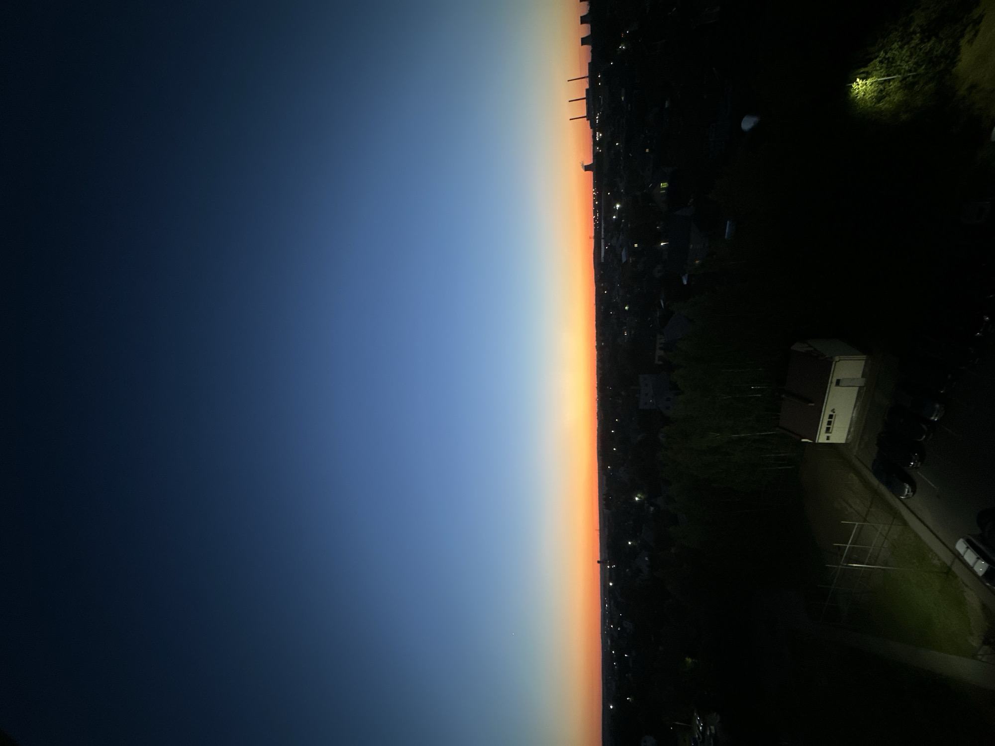 This is a high-angle, wide-shot photograph taken at dusk or dawn over the urban area of Schomyslitza, within the Schomyslitza rural council in Belarus. The sky dominates the upper portion of the image, transitioning from a deep navy blue at the zenith to a vibrant orange and yellow band just above the horizon. A single faint star is visible in the upper left quadrant of the sky.
Below the horizon, the silhouette of a city or town is visible. Numerous lights are scattered across the landscape, indicating a populated area. In the distance, to the right, several industrial structures, possibly power plant cooling towers and chimneys, are faintly illuminated against the colorful sky. The foreground of the image is much darker, showing a close-up view of a residential or recreational area.
In the lower left quadrant, there is a dark, grassy area with what appears to be a sports field, delineated by light-colored poles and fencing. Adjacent to this is a paved area with several parked cars, mostly dark in color, and one lighter-colored vehicle. To the right of the sports field, a small, light-colored building with a red roof is visible. Further to the right and in the background, more trees and buildings are obscured by the darkness. A solitary street lamp illuminates a patch of vegetation in the lower right corner. The scene is devoid of any visible people.