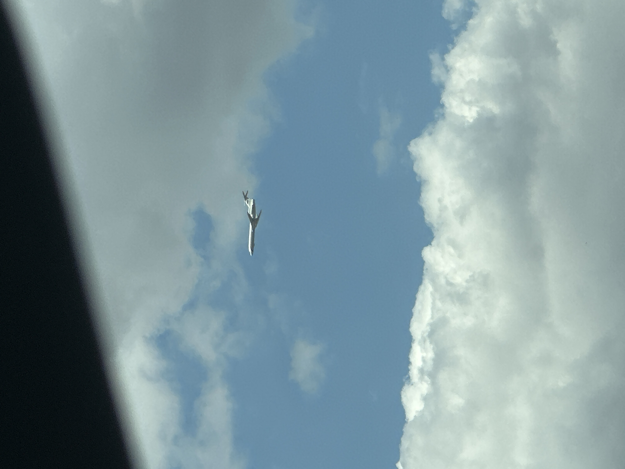 An airplane is visible in the sky, flying horizontally from left to right. The sky is blue with white clouds, and a dark shadow is cast across the top of the image, suggesting the photo was taken from inside a vehicle or from under an overhang. The airplane appears to be a commercial jet, white with two engines visible under the wings. It is positioned in the upper middle section of the frame. The overall scene depicts a clear day with some cloud cover. The location is specified as Bol'shoi Log, Russia.