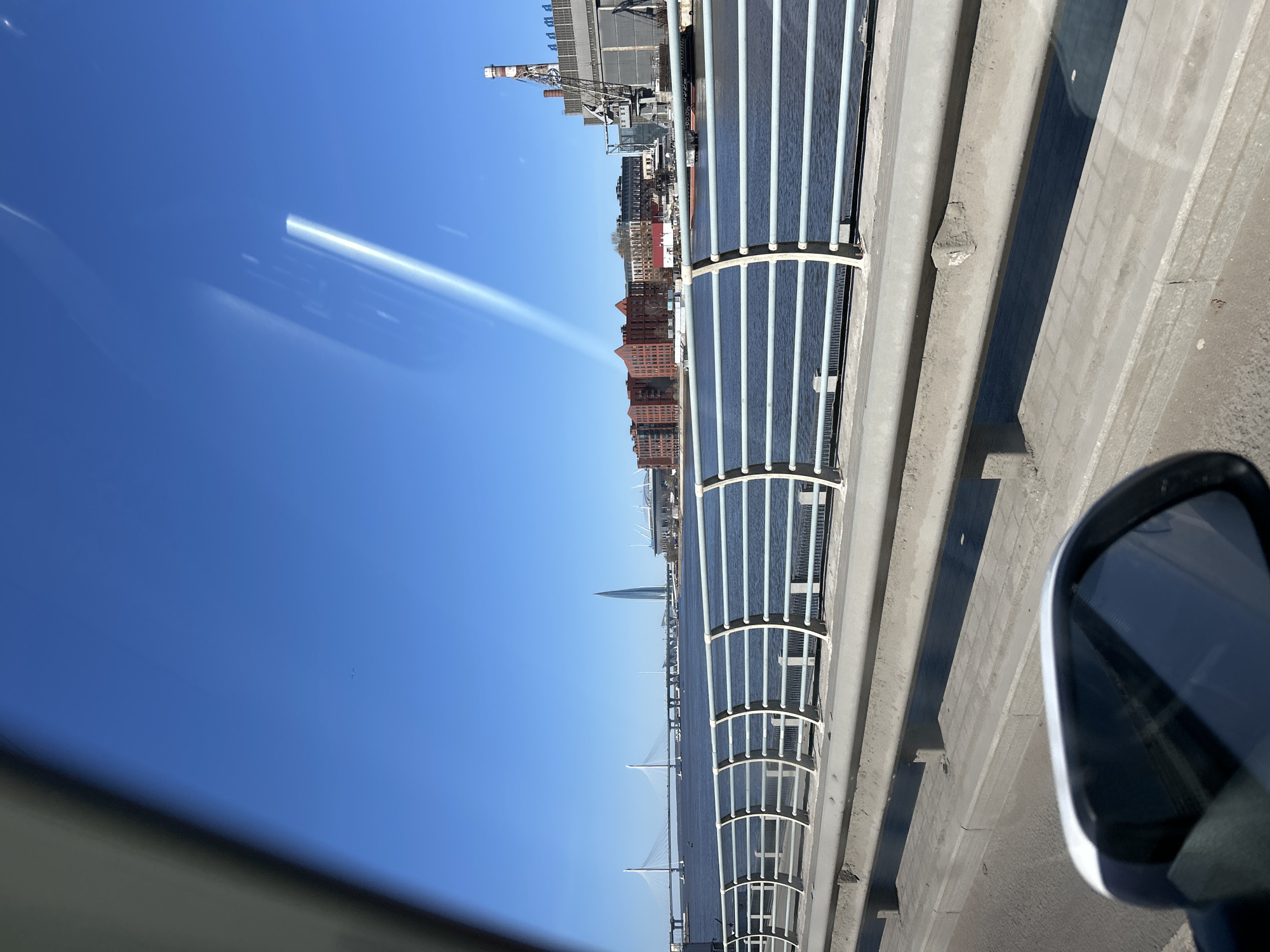 The image depicts a view from a vehicle, likely on a bridge, looking towards a cityscape under a clear blue sky. The bridge features a white-framed barrier running along its edge. Beyond the barrier, various buildings are visible, including what appears to be a skyscraper with a pointed top. To the left, a long bridge spans the water. A car side mirror is visible. The city is Saint Petersburg, Russia.