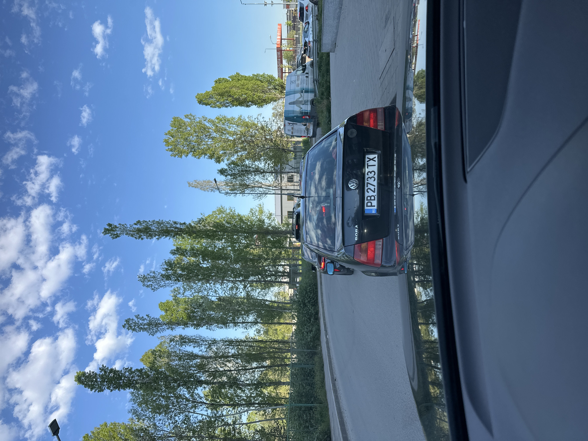 The image depicts a street scene in what appears to be a suburban or industrial area. The photograph was likely taken from inside a vehicle looking out to the right. The camera’s focus is primarily on a black VW Golf in the adjacent lane, which bears a Bulgarian license plate "PB 2733 TX". The view through the car window shows the edge of the car door and possibly the interior of the vehicle taking the image. The street features a line of tall, slender trees along the road. There is a building in the background and other parked vehicles. The sky is blue with scattered white clouds. Based on the license plate and the visual style, the location is highly probable to be somewhere in Sofia, Bulgaria.
