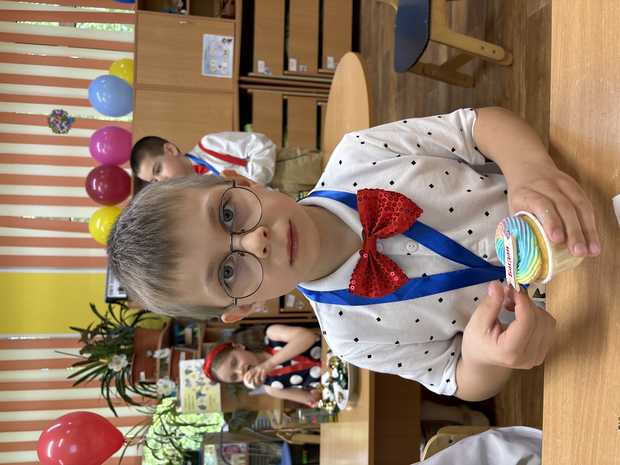 A young boy with short, gray hair, wearing round glasses, a white shirt with black dots, and a red sequined bow tie with a blue ribbon, sits at a wooden table, holding a cupcake with rainbow-colored frosting. The cupcake has a small tag with the name "Богдан" written on it. Behind him, another child is visible, wearing a white shirt with a red bow tie, along with other children in the background. Balloons of various colors (red, yellow, pink, and blue) are scattered throughout the room, and potted plants adorn the windowsill. A girl with a red headband is also present in the background, seated at another table. The image was captured indoors. The location details provided are Unknown, Unknown.