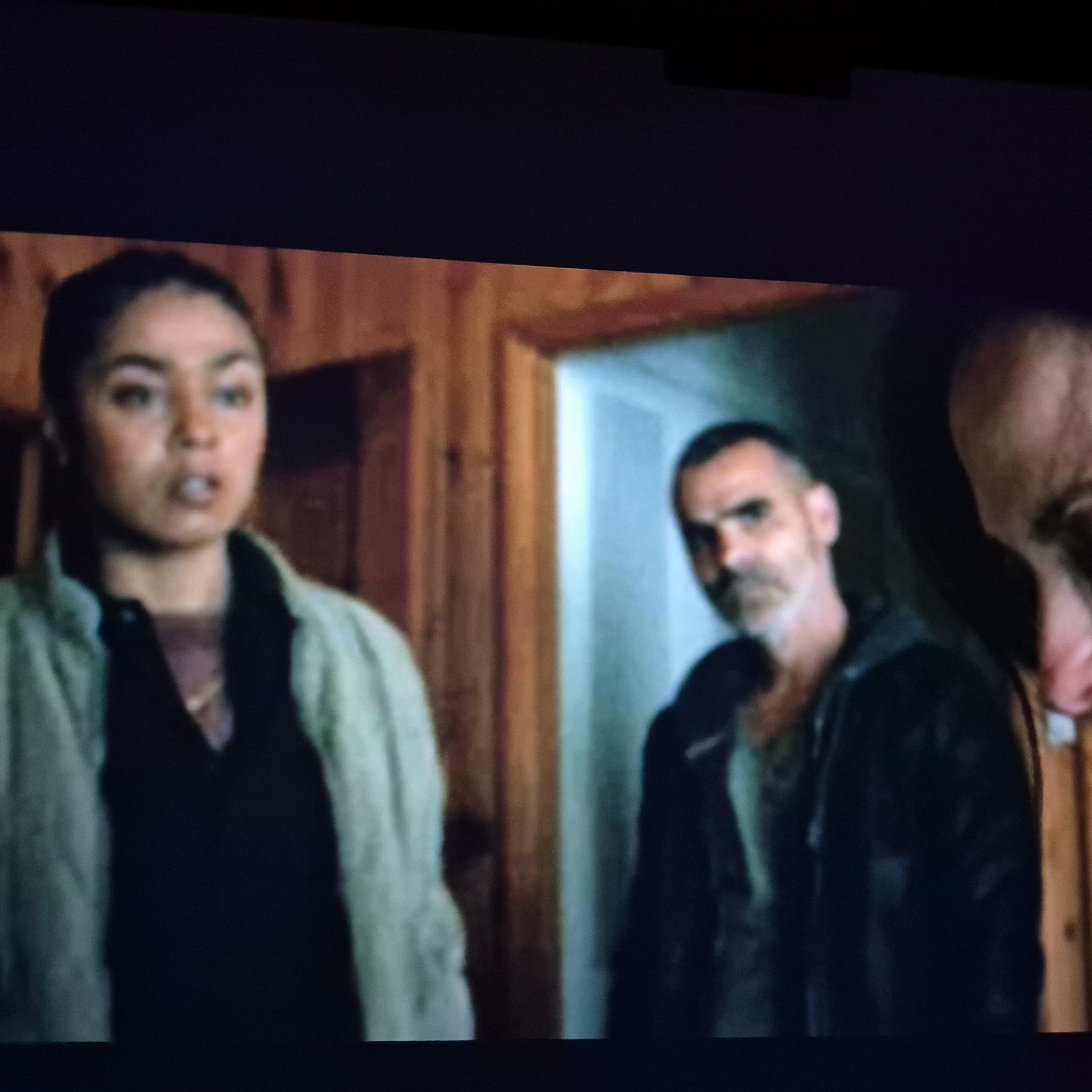 The image depicts a dimly lit interior scene with three individuals. In the foreground on the left is a woman with dark hair pulled back, wearing a light-colored jacket over a dark shirt. She is looking towards the viewer with an open mouth. To her right, a man with short, dark hair and a goatee is partially visible, wearing a dark jacket. Further to the right, another person's head and shoulder are visible, seemingly bald. The background consists of wooden paneling on the left and a lighter, possibly blue-toned wall or door on the right. The overall impression is that of a candid moment captured within a building, possibly a home or a similar indoor space. No specific details linking the scene to the city of Kępno, Poland, are discernible from the image itself.