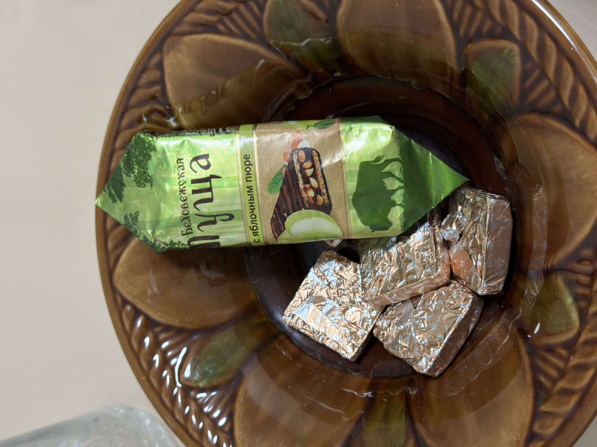 The image shows a brown, ornate ceramic bowl containing three individually wrapped candies in silver foil and one larger, unwrapped candy bar. The candy bar is wrapped in a green foil with text and images in Russian. The text on the candy bar reads "Беловежская Пуща" and "с яблочным пюре", which translates to "Belovezhskaya Pushcha" and "with apple puree". An image of a bison silhouette is visible at the bottom of the candy bar wrapper. The bowl is placed on a light-colored, possibly Formica countertop, and a portion of a clear drinking glass is visible on the left side of the frame. The scene depicts food items arranged in a bowl, suggesting a casual setting, likely a home or a cafe in Izhevsk, Russia. No individuals are present in the image.