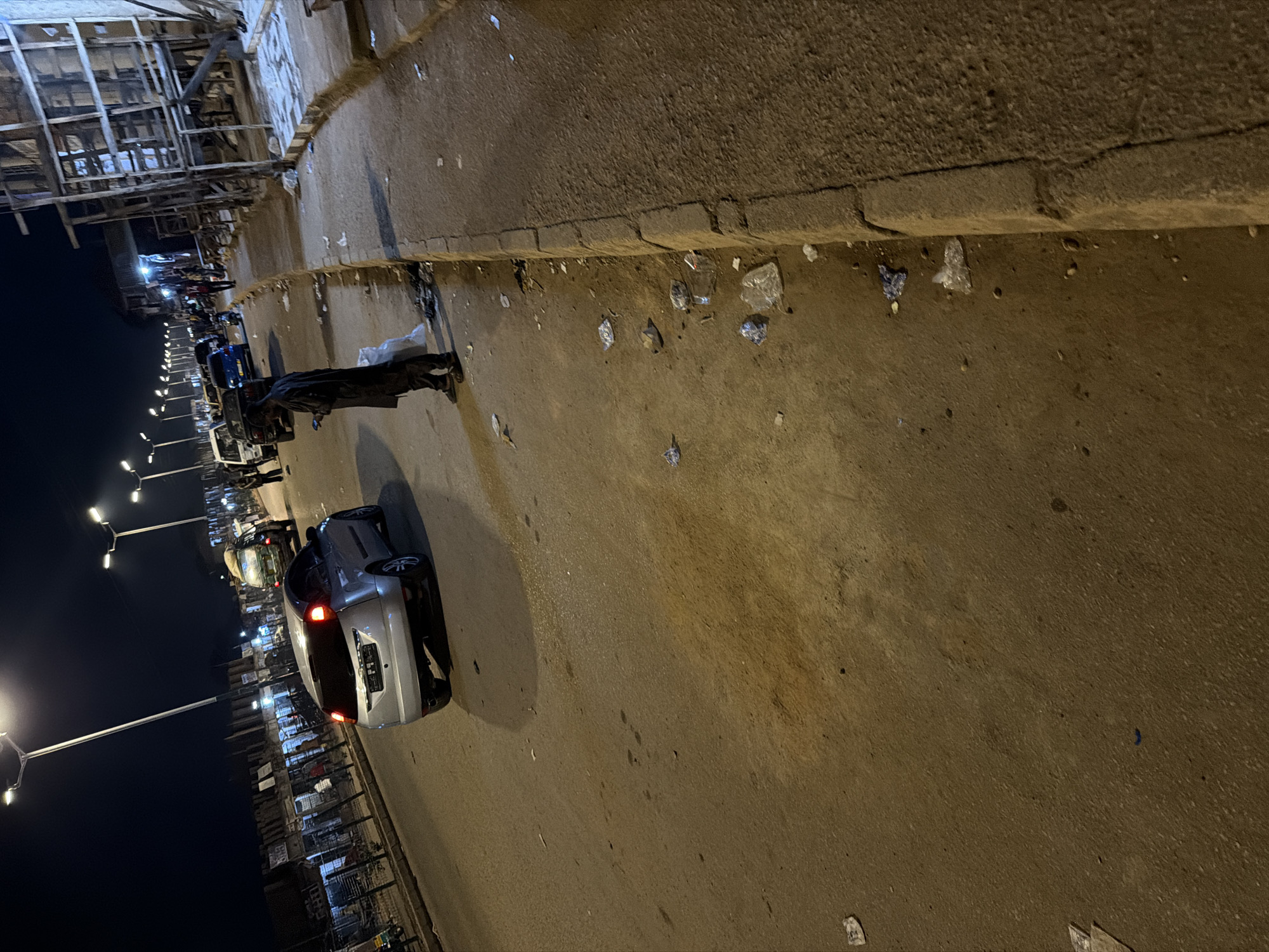 The image depicts a street scene at night in Bukuru, Nigeria. The foreground shows a paved road with scattered debris and a raised concrete curb along the right side. A silver car is positioned on the road, its brake lights illuminated. Further down the street, other vehicles, including a white van and a blue car, are visible. On the sidewalk to the left, a person wearing a dark traditional outfit is standing, looking at their phone. Beyond the vehicles, a row of structures, possibly shops or market stalls, line the street, illuminated by streetlights. The overall impression is one of a busy street at night, with evidence of everyday life and potential untidiness.
