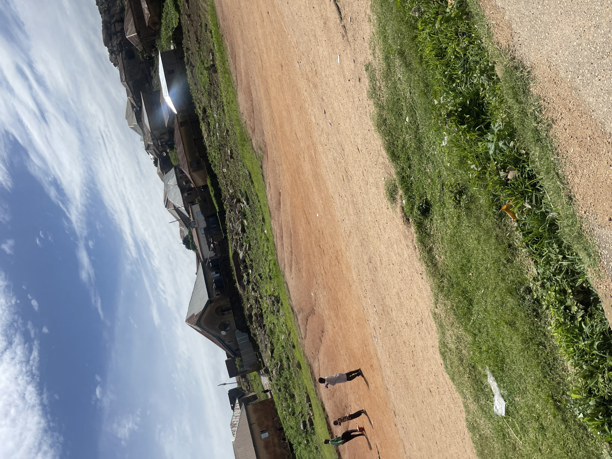 The image depicts a hilly residential area in Jos, Nigeria, under a partly cloudy sky with wispy clouds. In the foreground, a dusty, reddish-brown dirt path occupies the lower right quadrant, transitioning into a strip of green grass on the left. Three young boys are present on the dirt path. One boy, wearing a green long-sleeved shirt and black pants, appears to be kicking a soccer ball. Another boy, in a grey shirt and black pants, stands nearby, looking towards the first boy. A third, smaller boy in a white shirt and black pants, is also on the path.
Further up the slope, several houses with corrugated metal roofs are scattered across the hillside, built into the uneven terrain. Some of these houses are closer together, forming a cluster, while others are more spread out. The vegetation is sparse in some areas, with patches of green grass and rocky outcrops visible on the slopes. The overall impression is of a modest, developing community situated on a challenging, elevated landscape. The quality of the housing appears varied, with some structures appearing more weathered than others. The light suggests it is daytime.