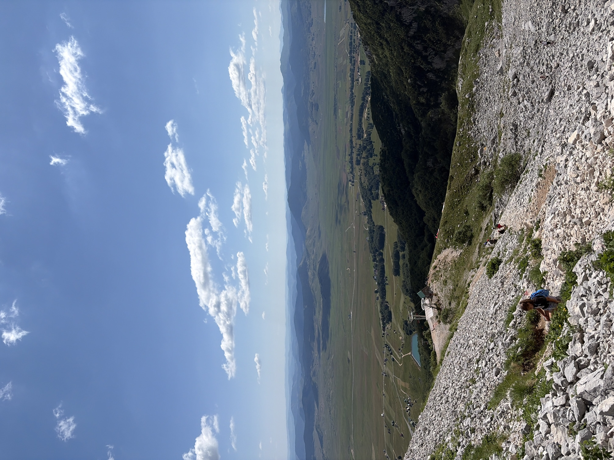 The image, taken in the vicinity of Pašina Voda, Montenegro, depicts a scenic mountain landscape under a bright blue sky with scattered white clouds. In the foreground, a steep, rocky slope descends, dotted with patches of green grass and small yellow flowers. A young person wearing a blue backpack is seated on the rocky terrain, interacting with the vegetation. Further down the slope, a dirt path winds its way, with several other individuals walking or resting.
A ski lift station with a visible pylon and gondola is situated on a lower plateau, adjacent to a small, turquoise body of water. Beyond this, the landscape opens into a vast, rolling valley. The valley floor is a mosaic of green fields, interspersed with trees and small clusters of houses, suggesting a rural settlement. A winding road is visible traversing the valley. In the background, successive layers of blue-green mountains recede into the distance, creating a sense of depth and grandeur. The overall scene conveys a feeling of outdoor recreation and the natural beauty of the Montenegrin highlands.