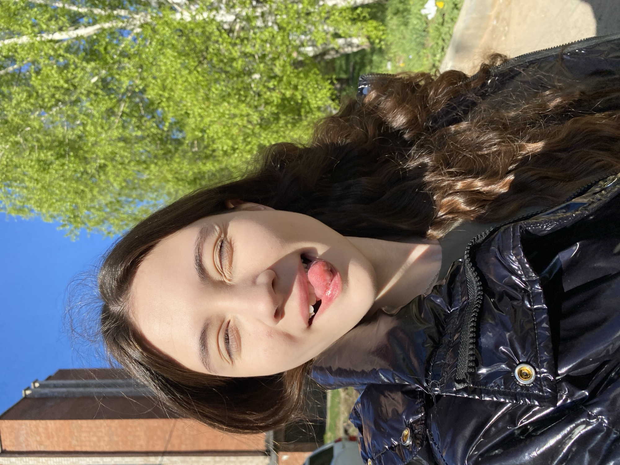 A young woman is outdoors in Votkinsk, Russia. She has long, dark hair with brown highlights and is wearing a shiny black puffer jacket. She is sticking her tongue out playfully. The background includes a brick building, a tree with bright green leaves, and a patch of sunlight. The image appears to be taken during the day.