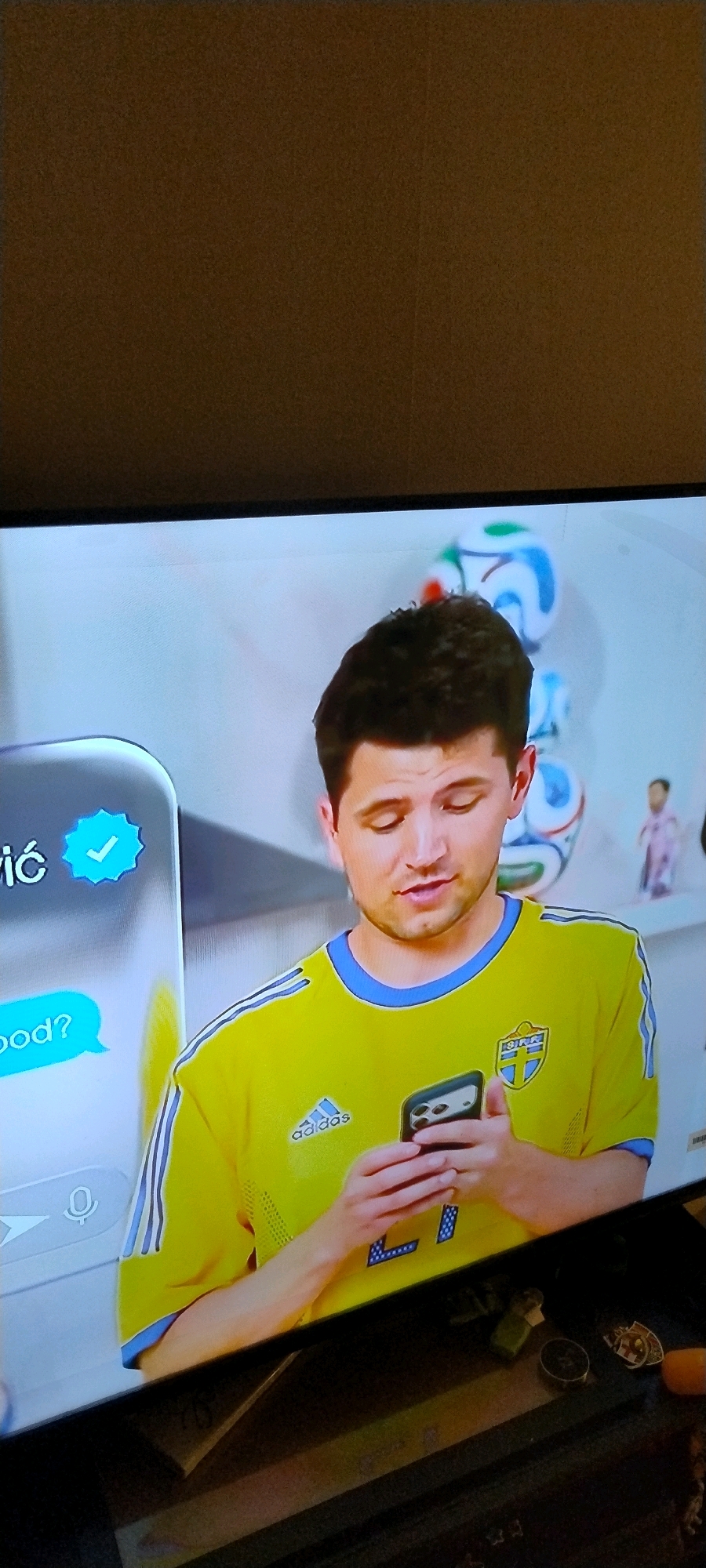 A young man with dark hair is seen on a television screen wearing a yellow and blue Adidas soccer jersey. He is holding a smartphone and looking down at it. To his left on the screen, there is a chat interface with text and a blue checkmark icon. In the background of the screen, there are several soccer balls and a small figurine of a person. The television is placed on a dark surface, possibly a TV stand or table, which is cluttered with various objects, including lighters, a circular object, and a medal. The scene appears to be indoors, with a neutral-colored wall visible behind the television. There is no information that places the image in Ishøj, Denmark.