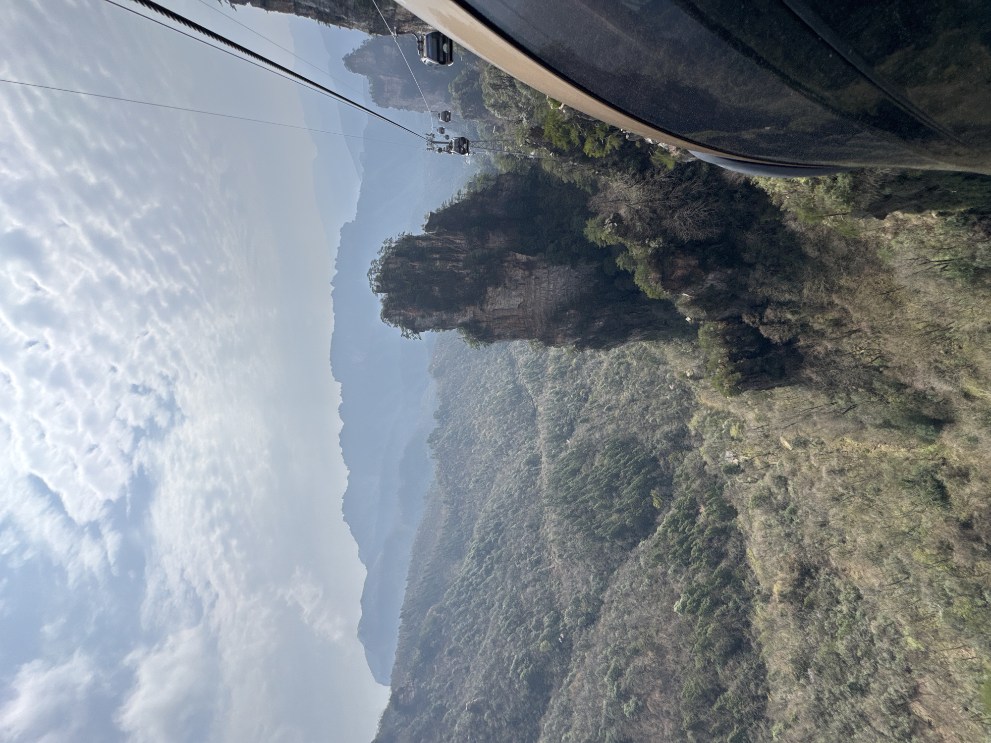 The image depicts a mountainous landscape in Wulingyuan, China, captured from the perspective of a moving cable car. The sky overhead is partially cloudy with a pattern of altocumulus-like clouds against a pale blue-grey expanse, indicating overcast or hazy conditions. In the midground, distinctive sandstone pillars, characteristic of Wulingyuan, rise steeply from the valley floor. One prominent, tall, and narrow karstic peak, topped with sparse coniferous trees, dominates the left-center of the frame. Surrounding this peak and extending across the valley are densely forested slopes and ravines, exhibiting a mix of deciduous trees with sparse foliage and some evergreen conifers, suggesting either late winter or early spring.
A cable car system is prominently featured, with multiple steel cables stretching across the scene. Several dark grey cable car gondolas are visible suspended from these cables in the mid-distance, traversing the mountainous terrain. A support pylon for the cable system is also discernible among the peaks. The right side of the image is occupied by the dark, metallic brown or bronze-colored exterior of the cable car cabin from which the photograph was taken, angled downwards and forward, reflecting some light. The overall scene conveys a vast, rugged natural environment adapted for human observation via aerial transport. No specific individuals are clearly identifiable, but the presence of the cable car system implies human activity and transit through the landscape.