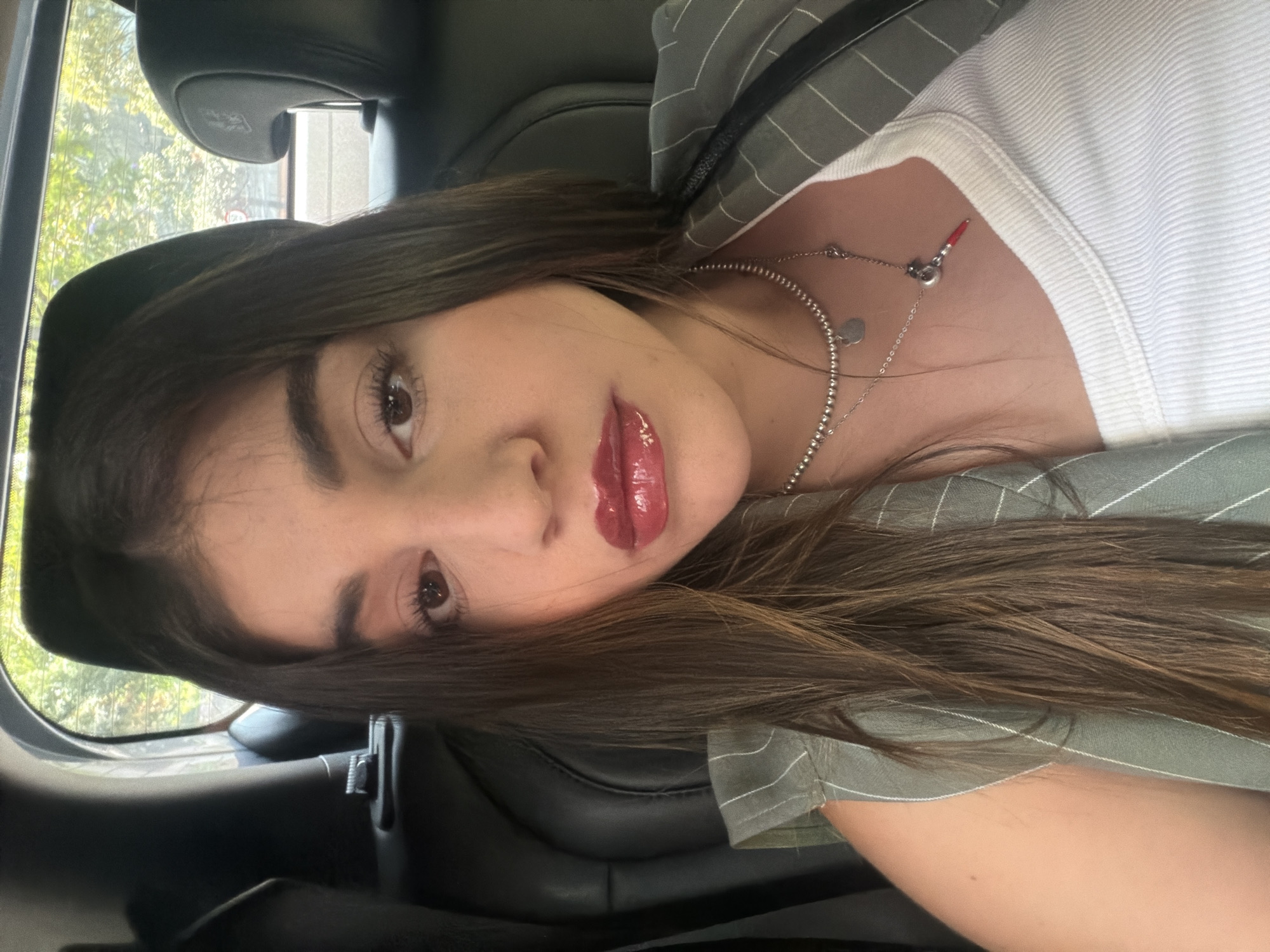 A young woman is taking a selfie in what appears to be the back seat of a car. The car is likely stationary. The location is unknown, but the presence of trees through the car window suggests the city of Mexico City, Mexico, is a possibility. The woman has long brown hair, brown eyes, and is wearing glossy red lipstick. She has on a white shirt and a striped jacket or blazer. Two silver necklaces adorn her neck. The car interior is black.