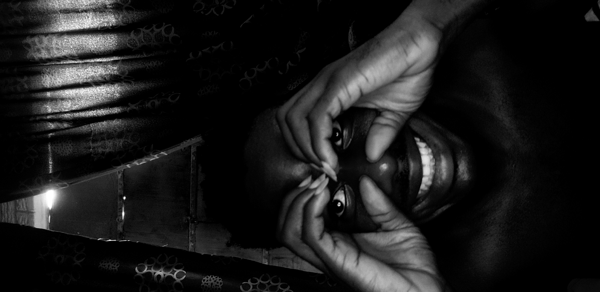 A monochrome, rotated image presents a close-up of a person with dark skin, exhibiting a joyful and playful expression. The individual's face is central, with both hands engaged in a dynamic gesture, seemingly pulling at the skin around their eyes and nose to exaggerate their features. A wide, beaming smile reveals a row of bright, prominent teeth. Their eyes are open wide and appear to be looking directly forward. The person's fingernails are visible and appear long, possibly manicured.
The background consists primarily of dark, patterned curtains that run across the top and bottom of the frame. These curtains have a textured or reflective quality, catching light in horizontal streaks. On the far left, a vertical streak of intense light is visible, suggesting an external light source or a gap in the curtains. The setting appears to be indoors, characterized by strong contrasts between deep shadows and bright highlights, enhanced by the black and white palette. The overall impression is one of lightheartedness and amusement. No visible text is present in the image.