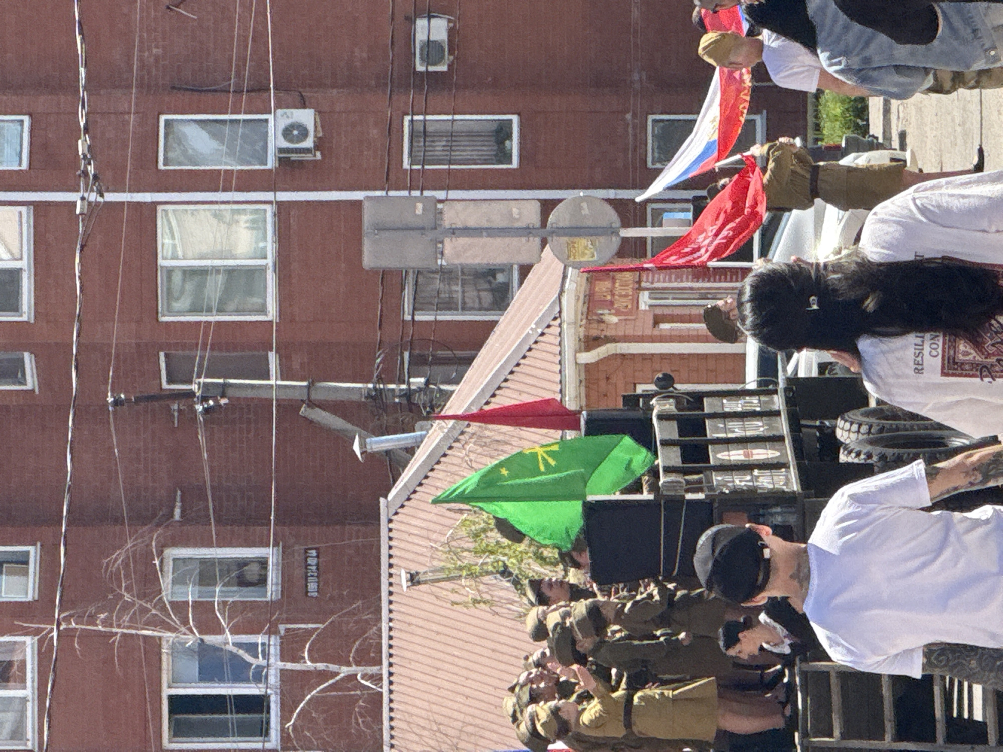 The image shows a procession in what appears to be an urban setting in Unknown, Unknown. A group of people dressed in military-style uniforms are visible, possibly veterans or participants in a commemorative event. They are on a vehicle adorned with flags, including a green one with a symbol and other red and Russian flags. The surrounding environment includes a red brick building with windows and an air conditioning unit attached to the wall. There are also utility poles and wires in the foreground. Several other people stand nearby, seemingly observing the procession. The overall impression is that of a public celebration or memorial event within the city.