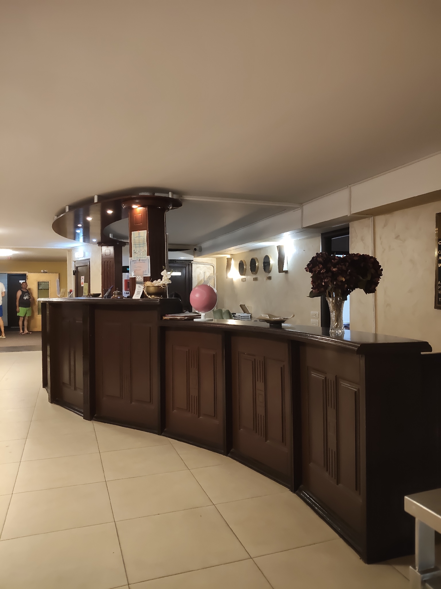 A wide shot captures the interior of a hotel lobby, featuring a prominent, dark wood reception desk that curves to the left. The desk has ornate paneling and is topped with various items. To the right, a crystal vase holds a large bouquet of dark red flowers. A pink globe on a stand sits to the far right of the desk. Behind the desk, in the center, a wooden pillar supports a curved structure extending overhead, adorned with spotlights. On the pillar, several documents are attached. To the left of the pillar, a small bell and a card reader are visible. Further left, a person wearing a black tank top and green shorts stands near a doorway, facing the desk. Another person, partially visible in a blue shirt and shorts, stands to the far left. The floor is tiled with large, light-colored square tiles. The walls are painted a neutral tone, with the wall behind the desk featuring a darker paneling or textured finish. A row of clock faces, representing different time zones, is mounted on the wall to the left of the flower arrangement. The ceiling is smooth and white, with recessed lighting. The overall lighting in the lobby is warm and inviting.