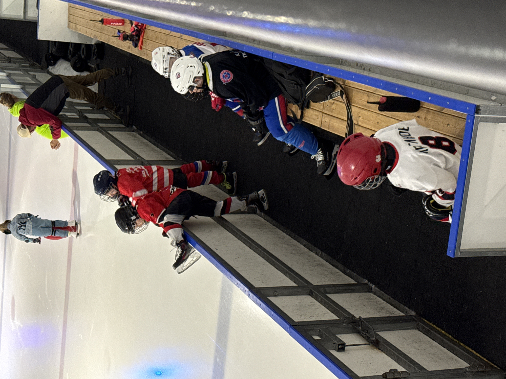 The image depicts an ice hockey rink with several young players and adults present. In the background, a young player in a denim outfit on skates is moving away from the camera. In the middle ground, a group of young hockey players in red and black uniforms are visible on the ice, with one player leaning over the boards. To the right, several young players are sitting on a wooden bench, wearing various colored helmets and uniforms, some with their skates on. Two adults are standing near the boards, observing the ice. The environment is an indoor ice rink with typical hockey rink features, including boards and protective netting. The lighting suggests an artificial indoor environment. The specific location in Votkinsk, Russia, cannot be determined from the visual information provided in the image.