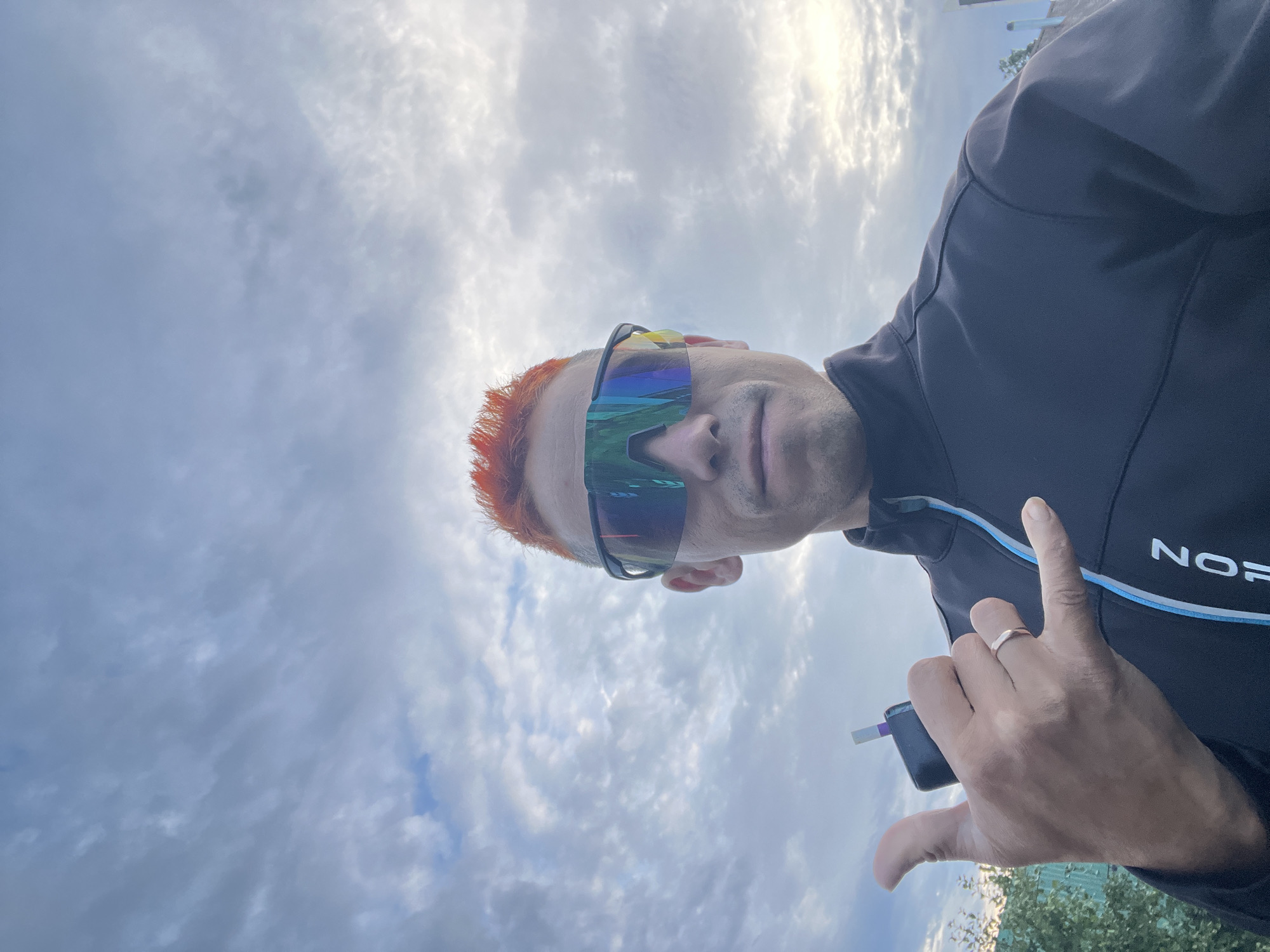 A man with bright orange, spiky hair is pictured outdoors against a cloudy sky. He is wearing a black jacket with blue and white trim and reflective, multi-colored sunglasses. In his left hand, he holds a black electronic device, possibly a vape, and is making a "shaka" hand gesture. A ring is visible on his left ring finger. The man appears to be in his 30s or 40s. The background shows a few green trees and hints of rooftops, suggesting an urban or suburban setting. The lighting indicates it is daytime, possibly late afternoon or early evening, given the soft light filtering through the clouds. The image is taken from a low angle, looking up at the man and the sky. The specific location in Chaikovsky, Russia is not discernible from the visual elements present in the photograph.