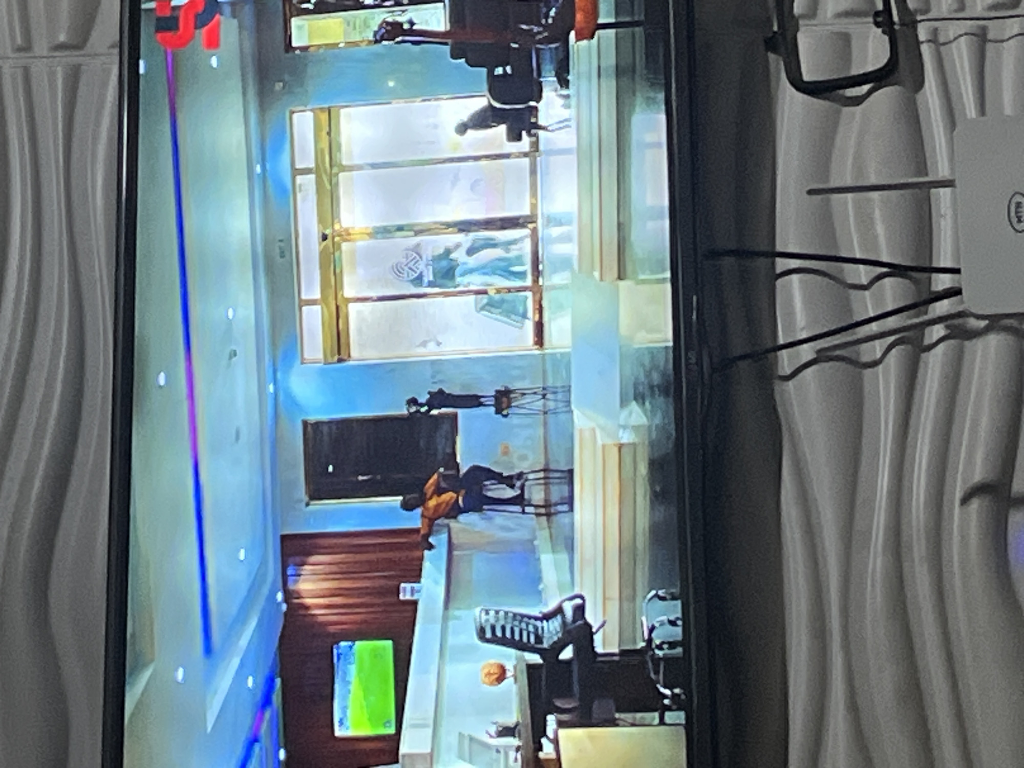 The image depicts a television screen displaying a scene that appears to be an indoor studio or reception area with modern architecture and lighting. In the foreground of the television screen, a person is seated on a tall stool at a counter, wearing an orange shirt and dark pants. To the left, a desk with an office chair is visible. A television screen displaying a green and blue image is mounted on the wall. In the background, automatic glass doors lead to an outdoor area, and several people are seen near the entrance. The overall ambiance suggests a contemporary commercial or entertainment setting. The lower portion of the image shows a textured wall and what appears to be a router and cables, indicating the television is in a room. The request to place this scene in Lagos, Nigeria is not supported by any visual cues within the image itself.