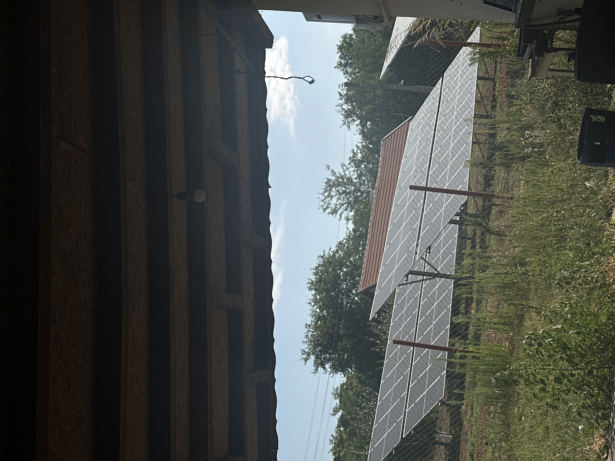 The image shows an outdoor scene with solar panels in the foreground and middle ground. The solar panels are arranged in rows and are tilted upwards, suggesting they are positioned to capture sunlight. In the background, there are trees and a roof of a building. The sky is visible above the trees, with some clouds. The overall environment appears to be rural, possibly a residential area or a farm, given the presence of solar energy infrastructure. The image seems to have been taken from under a covered structure, possibly a porch or a shed, as indicated by the wooden beams and ceiling in the upper part of the image. There is also some electrical wiring visible hanging from the ceiling. No people are visible in the image. The location is identified as Galiche, Bulgaria.