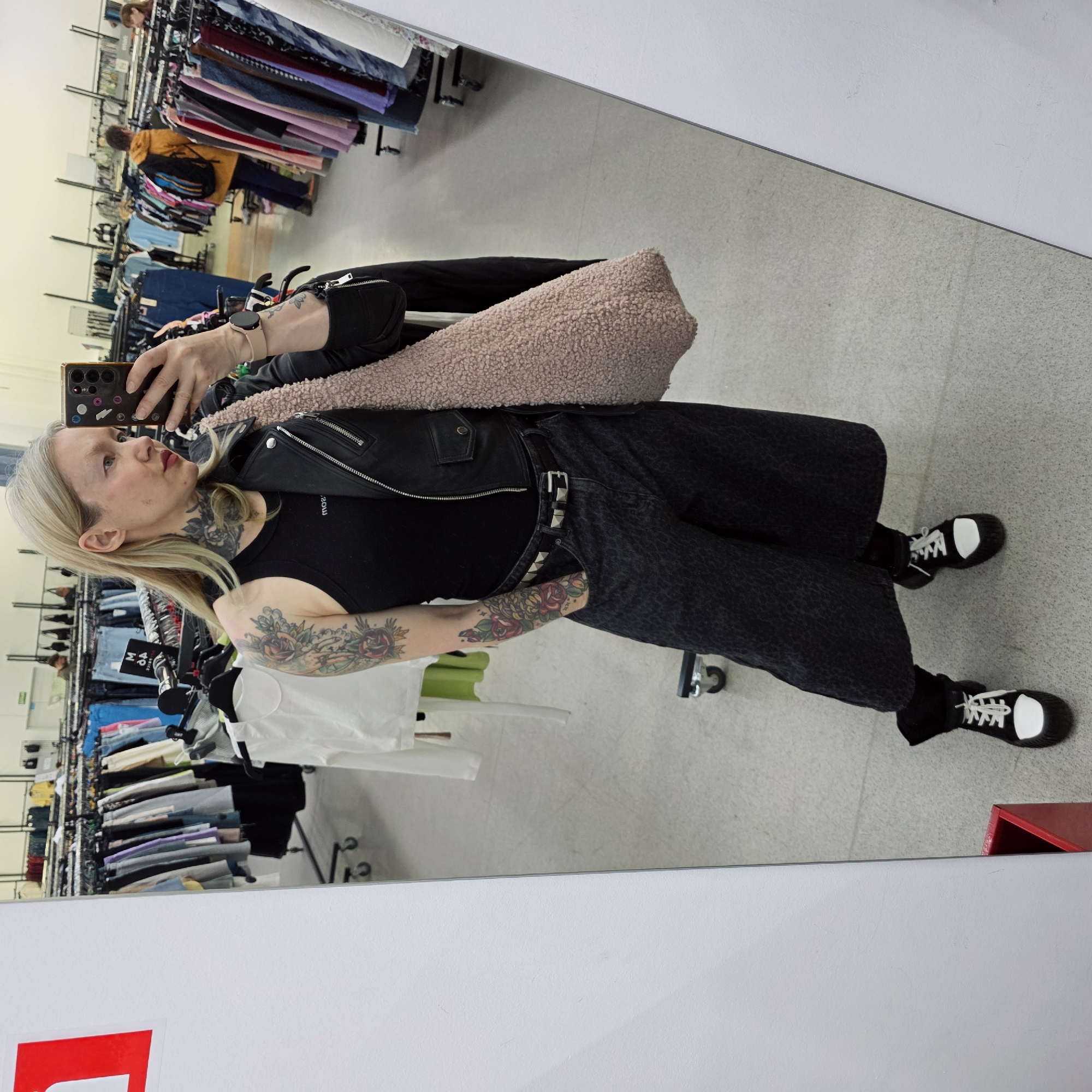 A person with blonde hair and multiple tattoos is taking a selfie in a mirror inside what appears to be a clothing store, likely a thrift store due to the extensive racks of used clothes in the background. The person is wearing a black tank top, a black leather jacket, black pants with a studded belt, and black sneakers with white toecaps. They have a large, tan, textured bag slung over their shoulder. The mirror reflects a long aisle of clothing racks filled with assorted garments. A red object is partially visible in the lower right corner of the image. The scene is well-lit. There is no information that indicates the location is Saint Petersburg, Russia.