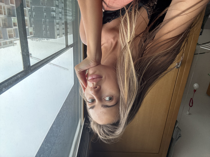 A female subject is depicted indoors, positioned to the right of a window or balcony door, looking directly at the viewer. She has long, straight light brown hair with blonde highlights, parted on the side. Her eyes are light-colored. Her right hand is positioned with fingers along her jawline, supporting her chin. She is wearing a black, lace-trimmed top.
The interior background on the left features a light-colored wall with a red-cabled ceiling light fixture holding a single bare incandescent bulb. Adjacent to this is a tall, light-brown wooden wardrobe or cabinet with a vertical handle. A peach-colored fabric object, likely a cushion or blanket, is partially visible in the lower right foreground.
Through the window, which exhibits visible smudges, a modern urban landscape unfolds under an overcast sky. Immediately outside, a dark-colored balcony railing with multiple horizontal bars is present. Beyond the railing, a series of multi-story residential buildings with grey facades and contemporary, modular designs are visible, some appearing to be in various stages of construction or recent completion. Two distinct tower cranes are prominent in the middle distance: one is a luffing-jib crane, and another, a hammerhead crane, indicating significant ongoing urban development. This architectural style and extensive construction activity are characteristic of rapidly expanding districts within Warsaw, Poland, such as Wola or Ochota.