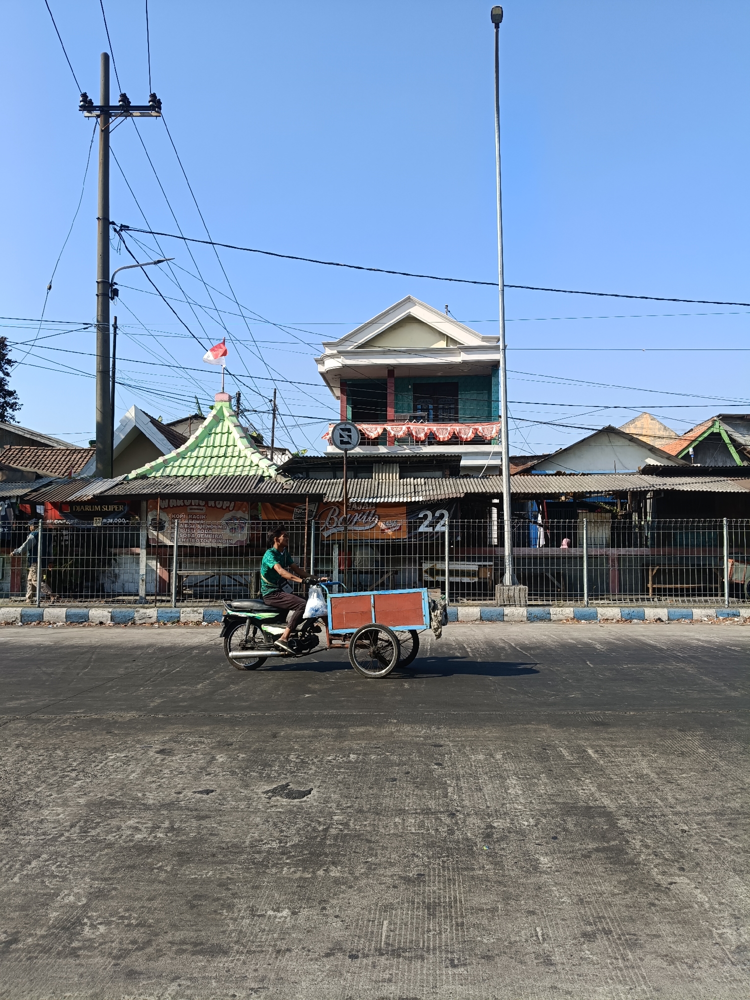 An outdoor, eye-level, full shot captures a man riding a motorcycle with a sidecar down a paved street in Kedungrejo, Indonesia. The sky is clear and blue. The man, wearing a green shirt and black pants, is positioned on the motorcycle. The motorcycle is equipped with a red and blue sidecar that is empty. Behind the motorcycle, a metal fence separates the street from a row of buildings. The buildings appear to be shops or residences with various roof styles, including a distinctive green, multi-tiered roof topped with an Indonesian flag. Power lines are strung across the scene, supported by a tall, grey utility pole on the left. A tall street lamp stands on the right. The street itself is dark asphalt with some wear and tear. The overall impression is a typical street scene in an Indonesian town.