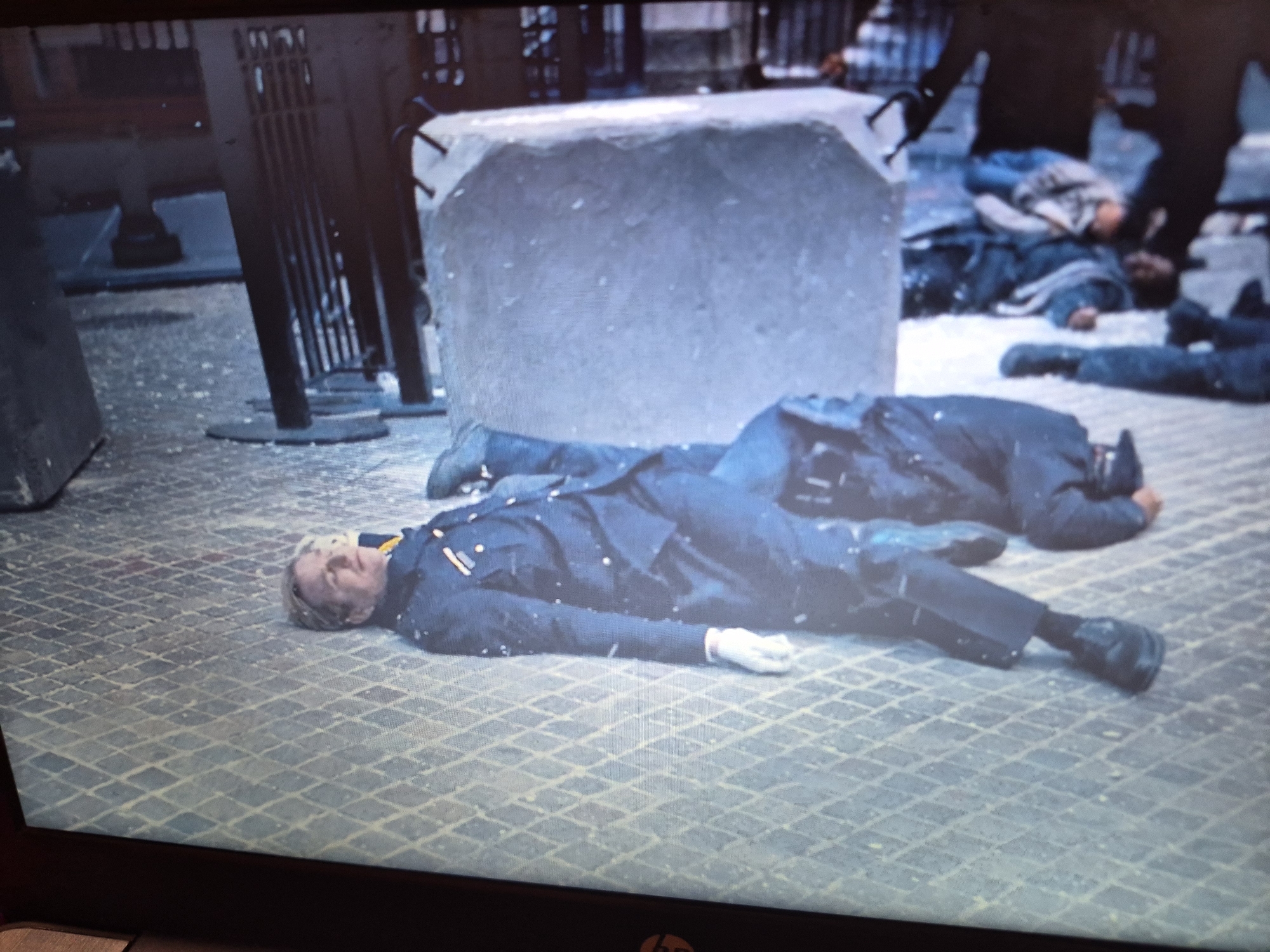 A man in a dark blue uniform, possibly a police or military uniform, lies on his back on a paved surface that appears to be made of diamond-shaped stones. He is wearing a white glove on his right hand, which is extended forward. His eyes are closed, and his head is tilted back. Snow or light debris is visible on his uniform and the ground. Behind him, another person in a dark uniform is lying on their side, partially obscured. In the background, a large, rough, light-colored object, possibly concrete or stone, is visible. There are other figures in the background who appear to be lying on the ground. The overall scene suggests a depiction of a historical event or a staged scene, possibly a memorial or reenactment, given the artificial appearance of the large object. The location is not identifiable as Sandwell, United Kingdom from the visual information present.