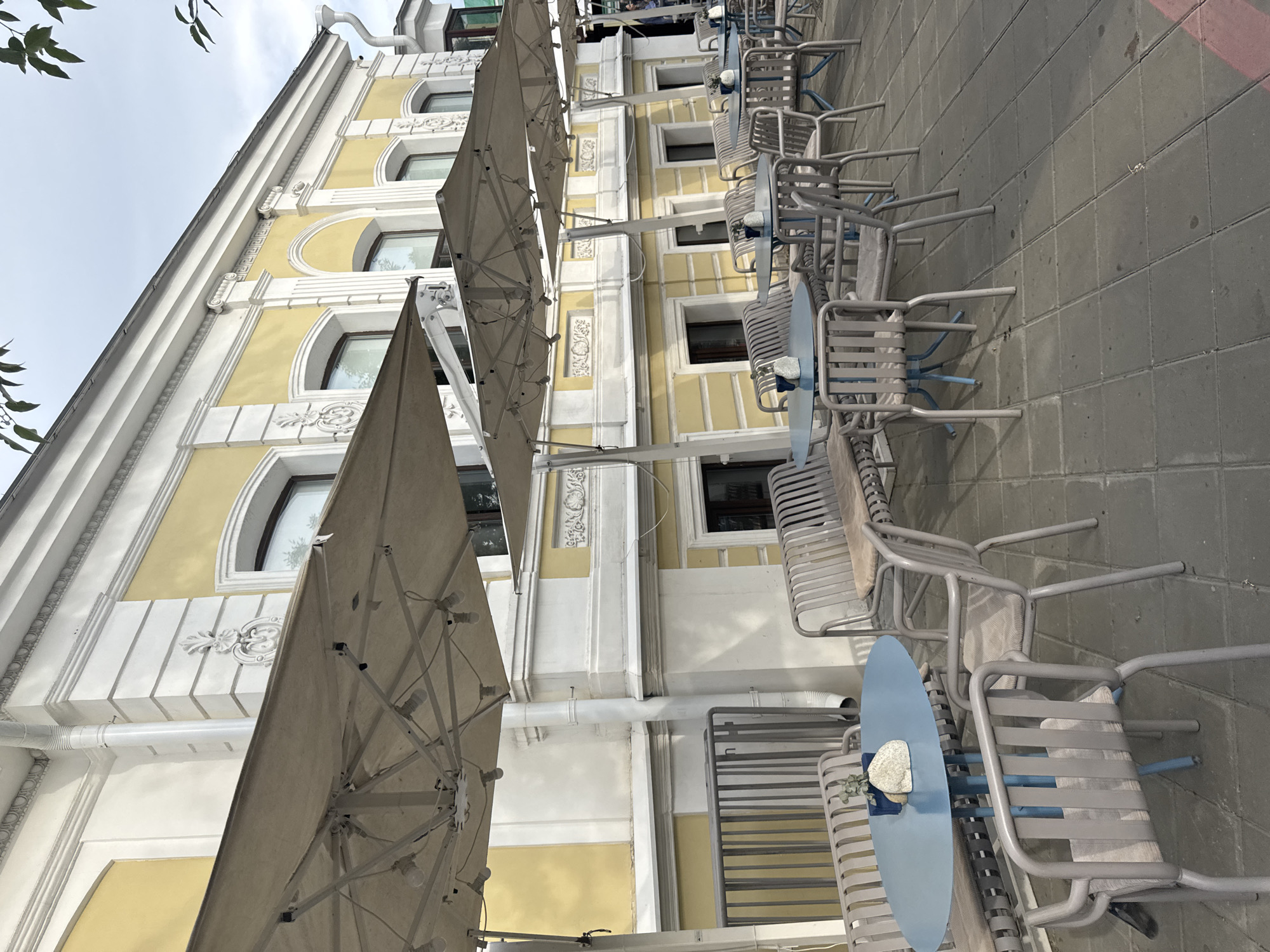The image shows an outdoor seating area of a cafe or restaurant on a cobblestone patio next to a building. The building is tall and painted a pale yellow with white decorative trim around the arched windows and along the facade. Several large, light-colored umbrellas are extended over the seating area. Underneath the umbrellas, there are multiple tables and chairs. The tables appear to be round and light blue. The chairs are made of metal with a light grey frame and light-colored woven seats and backs. Some of the chairs are empty, while others are occupied by people. The people are casually dressed and appear to be enjoying the outdoor atmosphere. The sky is overcast, suggesting it might be a cloudy day. The scene is set in Yekaterinburg, Russia. There is no visible text in the image.