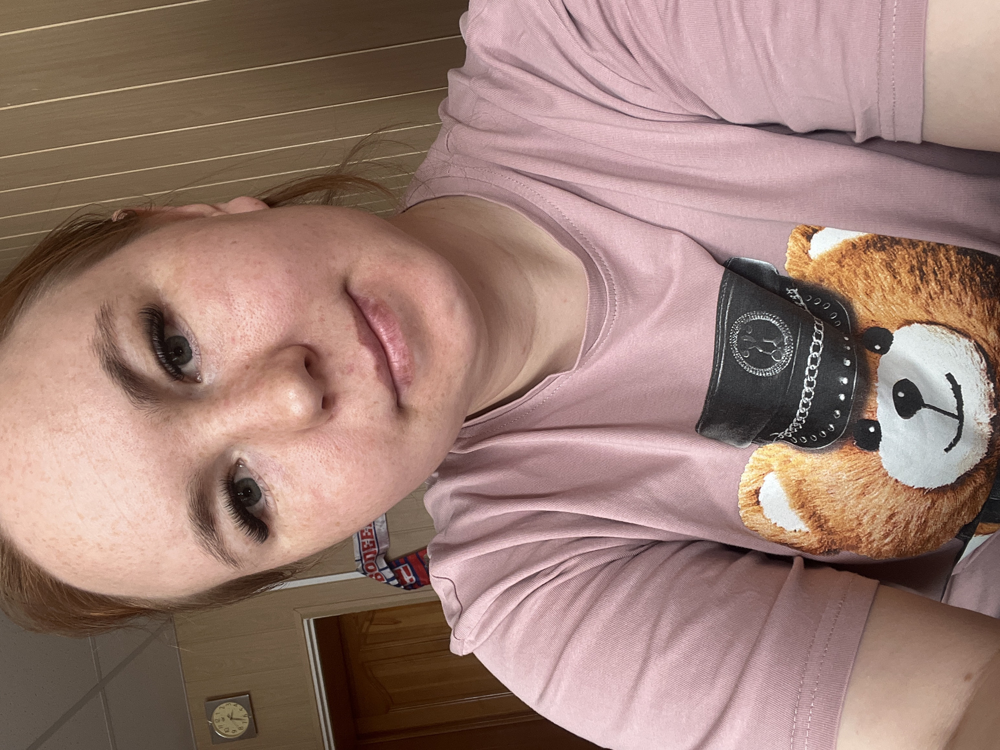 A light-skinned woman with freckles is the primary subject of this close-up photograph. She is wearing a light pink t-shirt featuring a print of a teddy bear wearing a black leather cap. The woman is looking directly into the camera. The photo is taken indoors, with a light brown wooden wall and doorway visible in the background. A clock is visible in the lower left corner of the photograph. A portion of red and blue patterned fabric is visible near the woman's arm. No specific location in Chaikovsky, Russia is identifiable from this image.