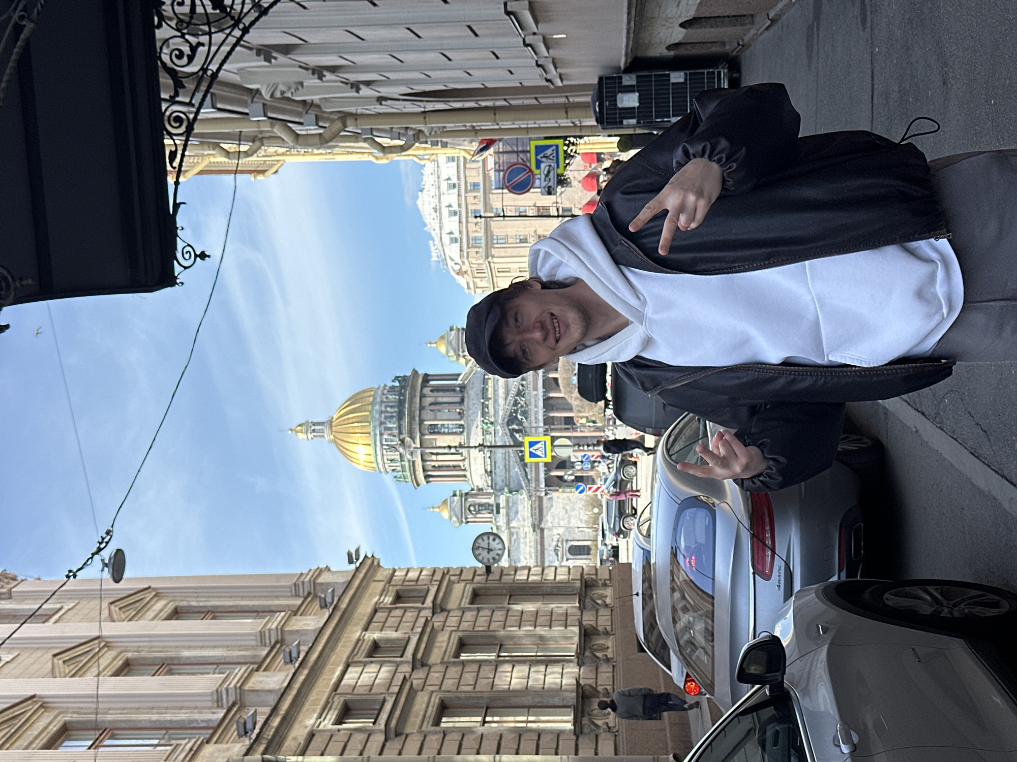 The image depicts a vibrant street scene in Saint Petersburg, Russia, centered around a man posing happily with St. Isaac's Cathedral visible in the background. The man, positioned to the right of the frame, is smiling broadly and making peace signs with both hands. He wears a black baseball cap, a white hooded sweatshirt, a black jacket, and grey pants.
Behind the man, the grand golden dome and classical architecture of St. Isaac's Cathedral stand prominently under a clear, bright blue sky, suggesting a sunny daytime. The street is lined with historic buildings featuring ornate facades and decorative elements, including a black wrought-iron awning in the foreground.
Several cars are parked along the street on the right, including a silver sedan displaying the "4MATIC" emblem on its rear. Various road signs are visible, such as a blue pedestrian crossing sign and a round blue sign with a red diagonal line (indicating "No Stopping"). A small clock is mounted on a building facade in the mid-ground. The scene conveys a lively urban atmosphere with historical significance.