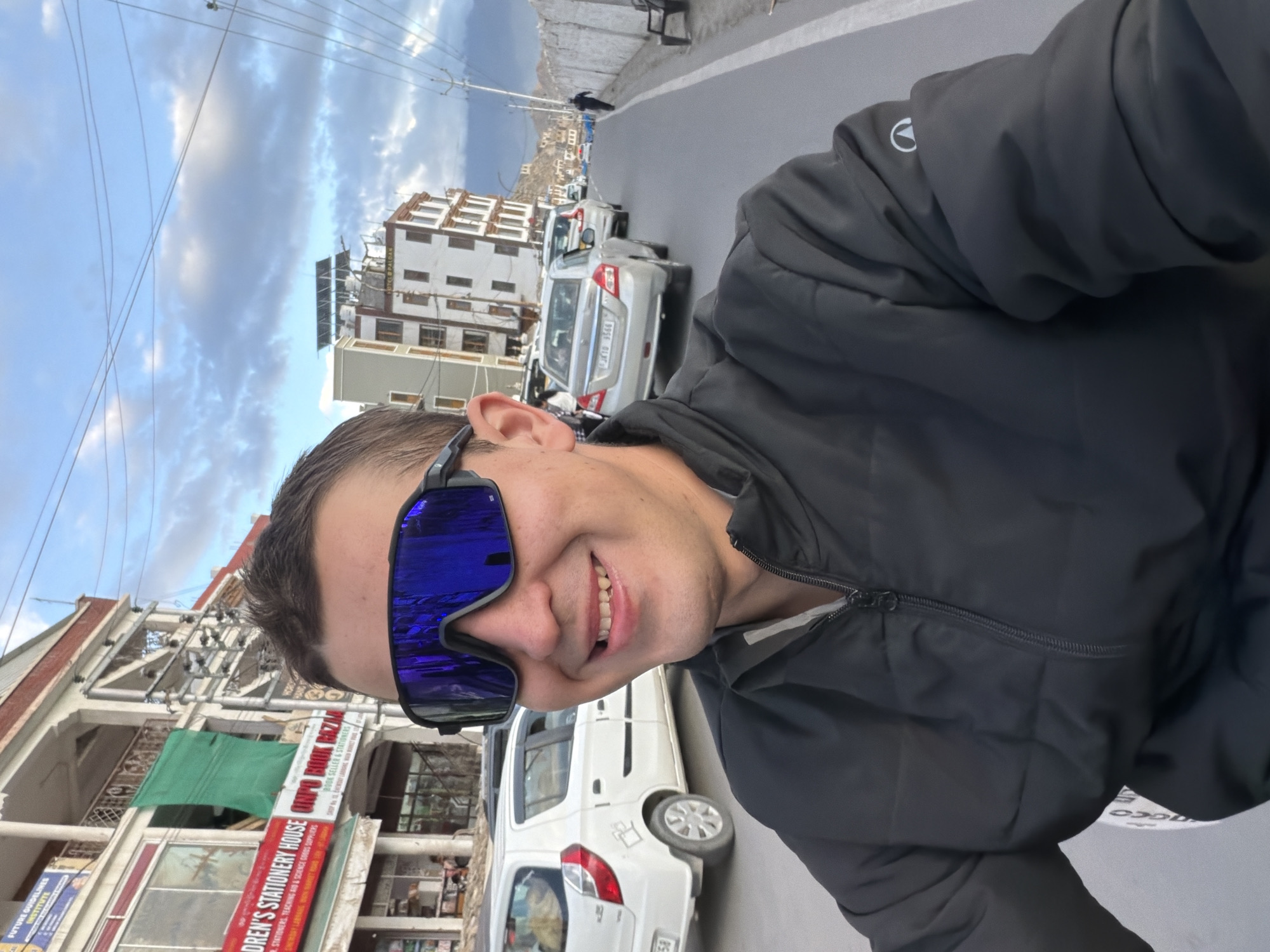 In Leh, India, a man with short brown hair, wearing large blue reflective sunglasses and a black zippered jacket, smiles directly at the camera. He is on a street with several cars driving in the same direction. To his left, buildings line the street, with signs indicating businesses like "Children's Stationery House" and "Ungo Book Bazaar." The sky above is partly cloudy with a mix of blue and gray clouds. The road is paved, and a white wall runs along the right side of the street, with a bench in front of it. The overall scene suggests a bright, daytime setting in a bustling urban environment.