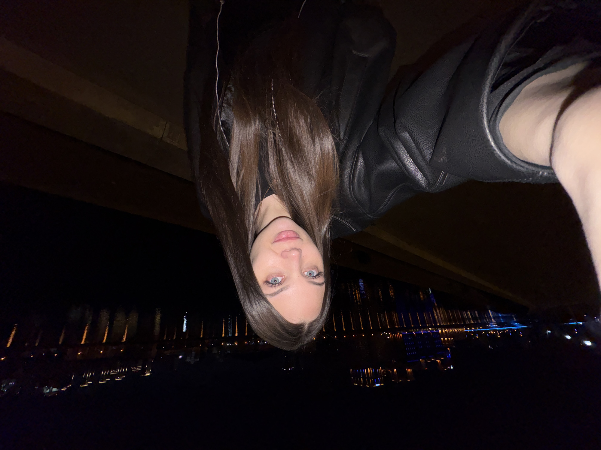 A woman is pictured against a dark cityscape, likely at night. She has light eyes, dark hair and is wearing a black jacket. The cityscape appears to be reflected in water, with scattered lights. The photo seems to be taken from an elevated position. The image does not contain enough detail to identify a specific location in Yekaterinburg.